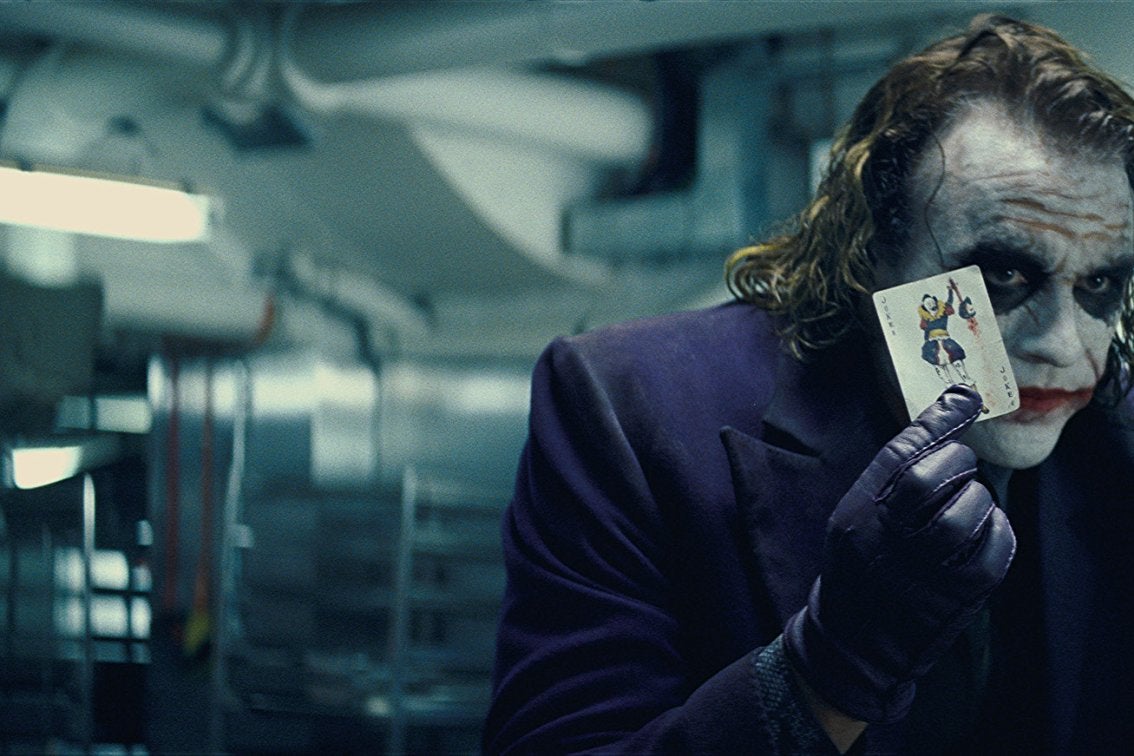 Heath Ledger as the Joker in The Dark Knight. He wears smeared, clown-like makeup and holds a playing card.