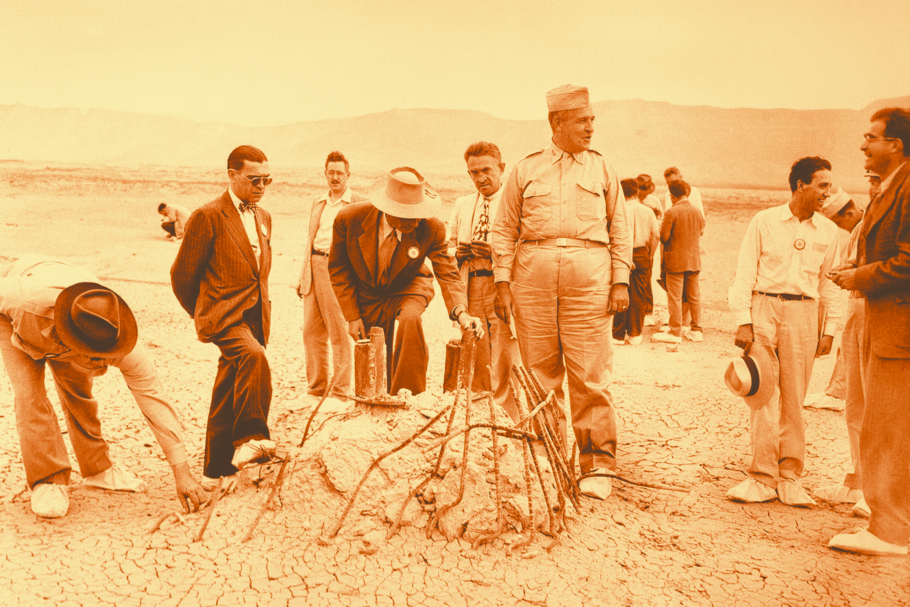 A black and white photo, tinted orange, of a group of men in suits and a general in uniform gathered around twisted rebar protruding from dry, cracked sand in the desert.