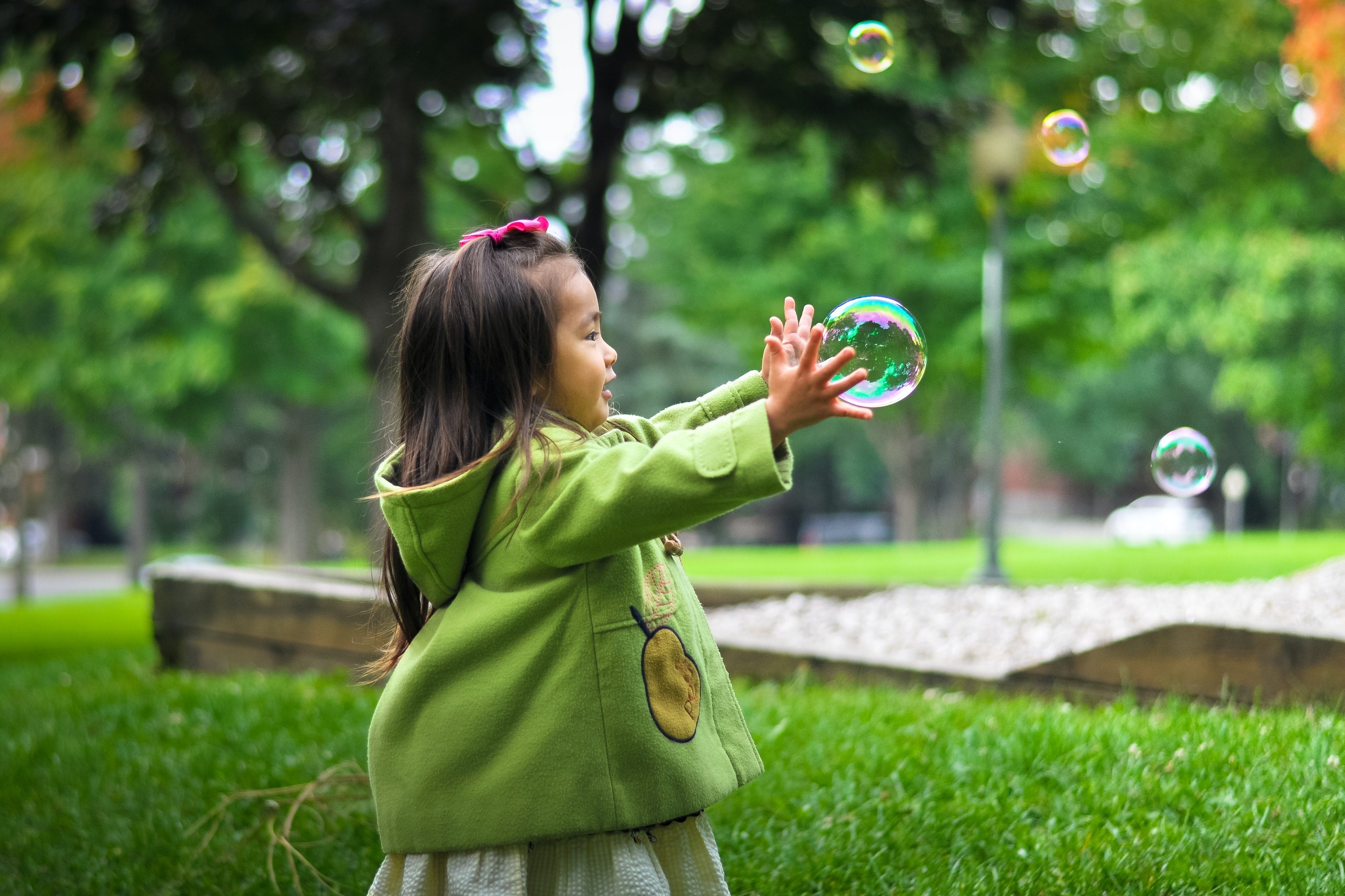 A little girl in a park-like setting reaches for bubbles.