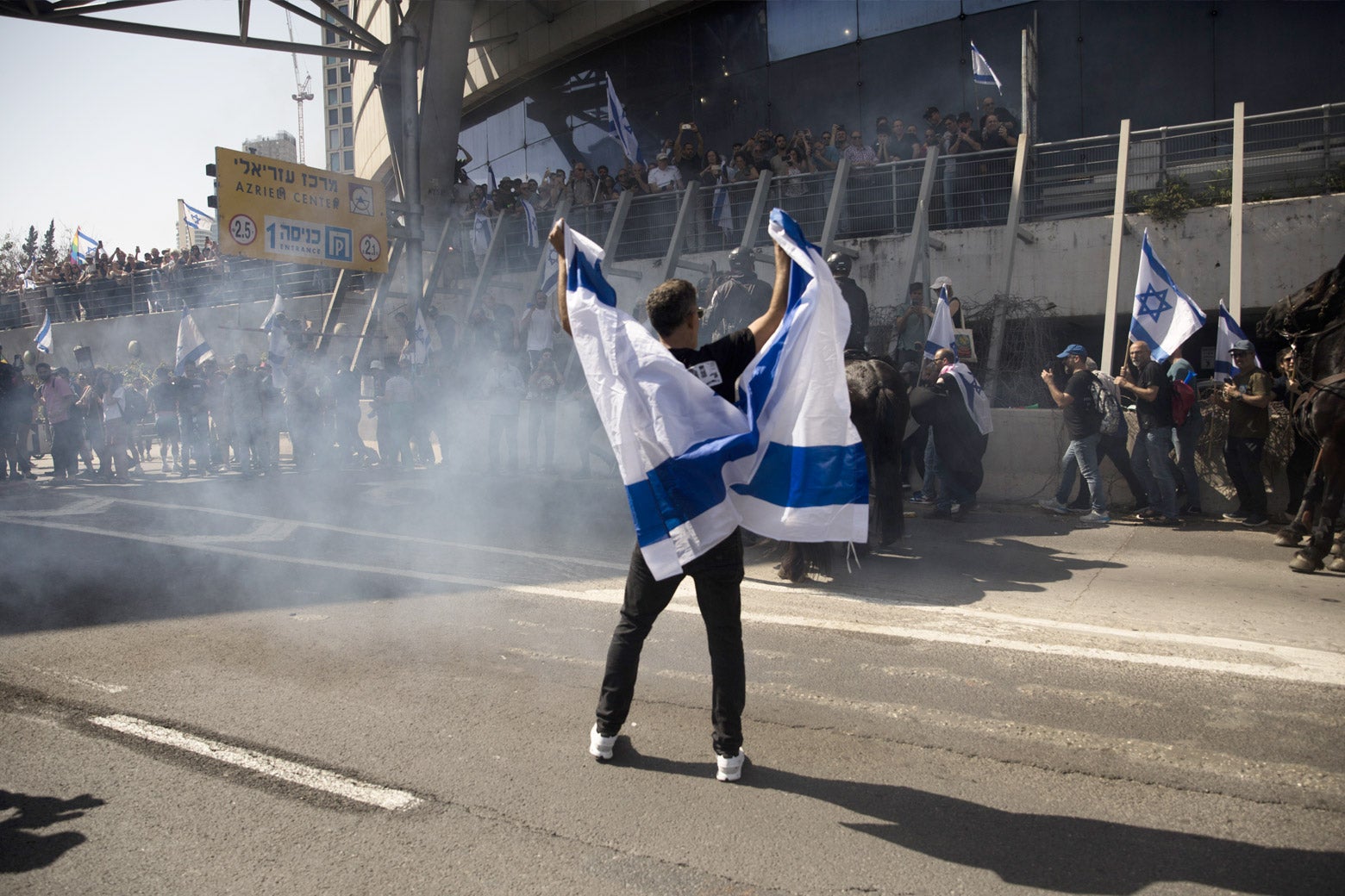 An Israeli protester waves the Israeli flag during clashes with Israeli police officers in a demonstration against the Israeli government's judicial reform plan on March 1, 2023 in Tel Aviv, Israel. Smoke rises around him.