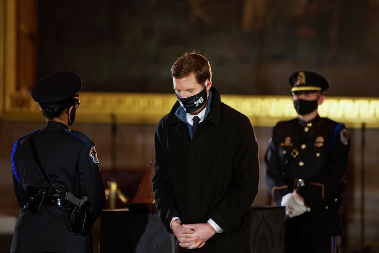 Lamb stands with his head bowed and hands clasped in front of him. One uniformed officer stands in front of him and one behind him.