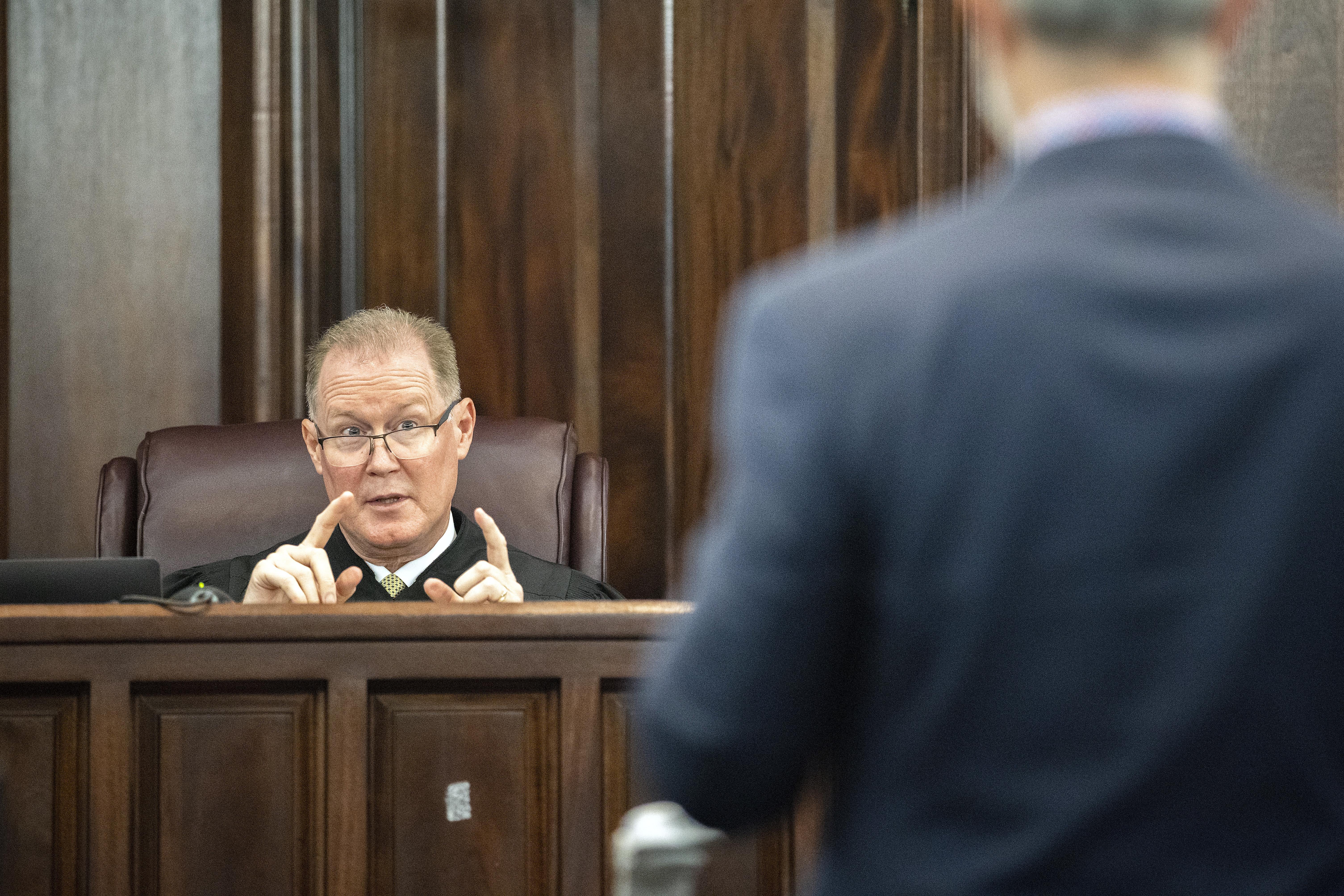 Judge sitting on the bench gestures as he speaks to a man standing in the foreground