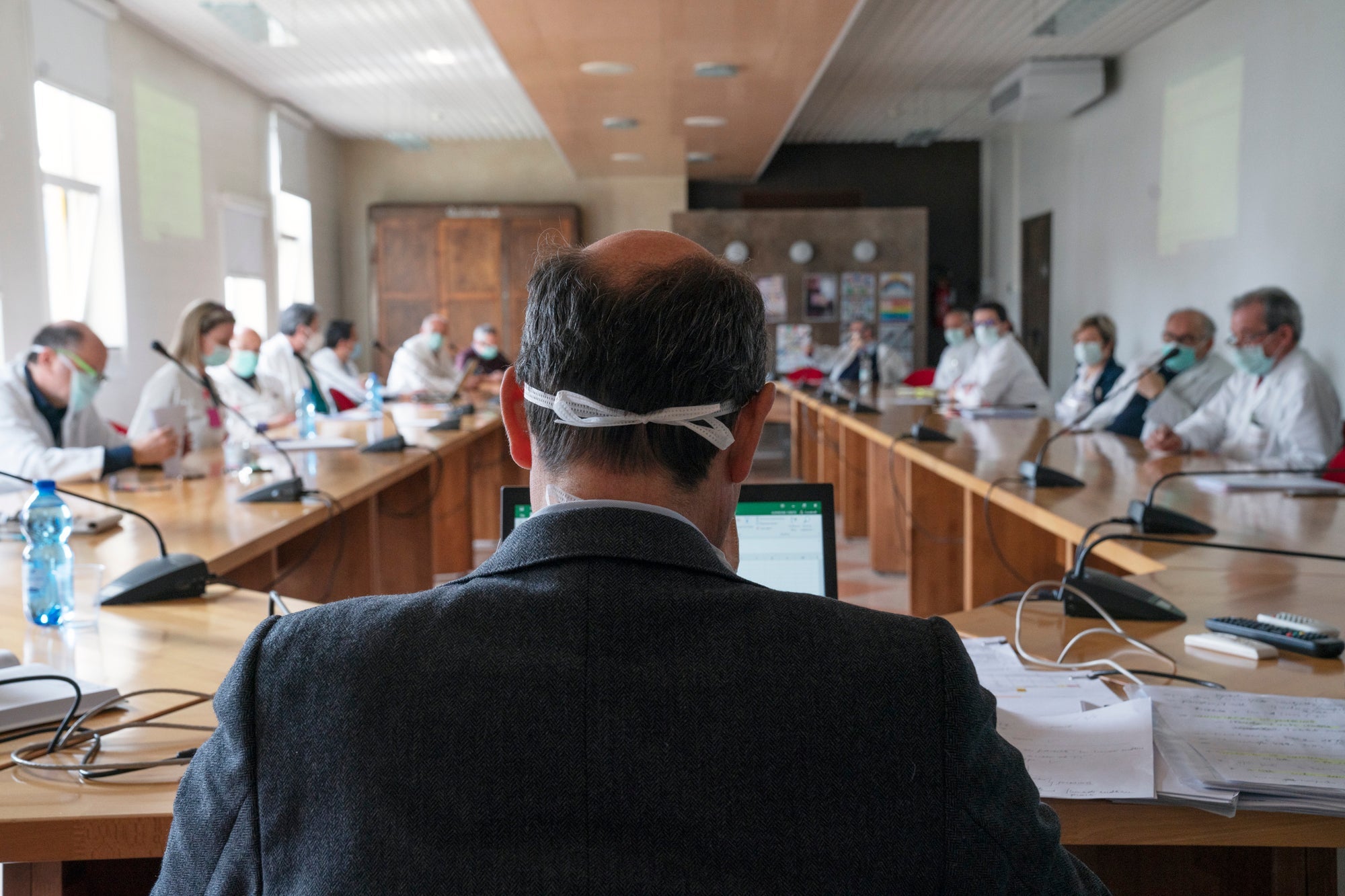 A man in a suit wearing a surgical mask briefs a conference room full of masked doctors.