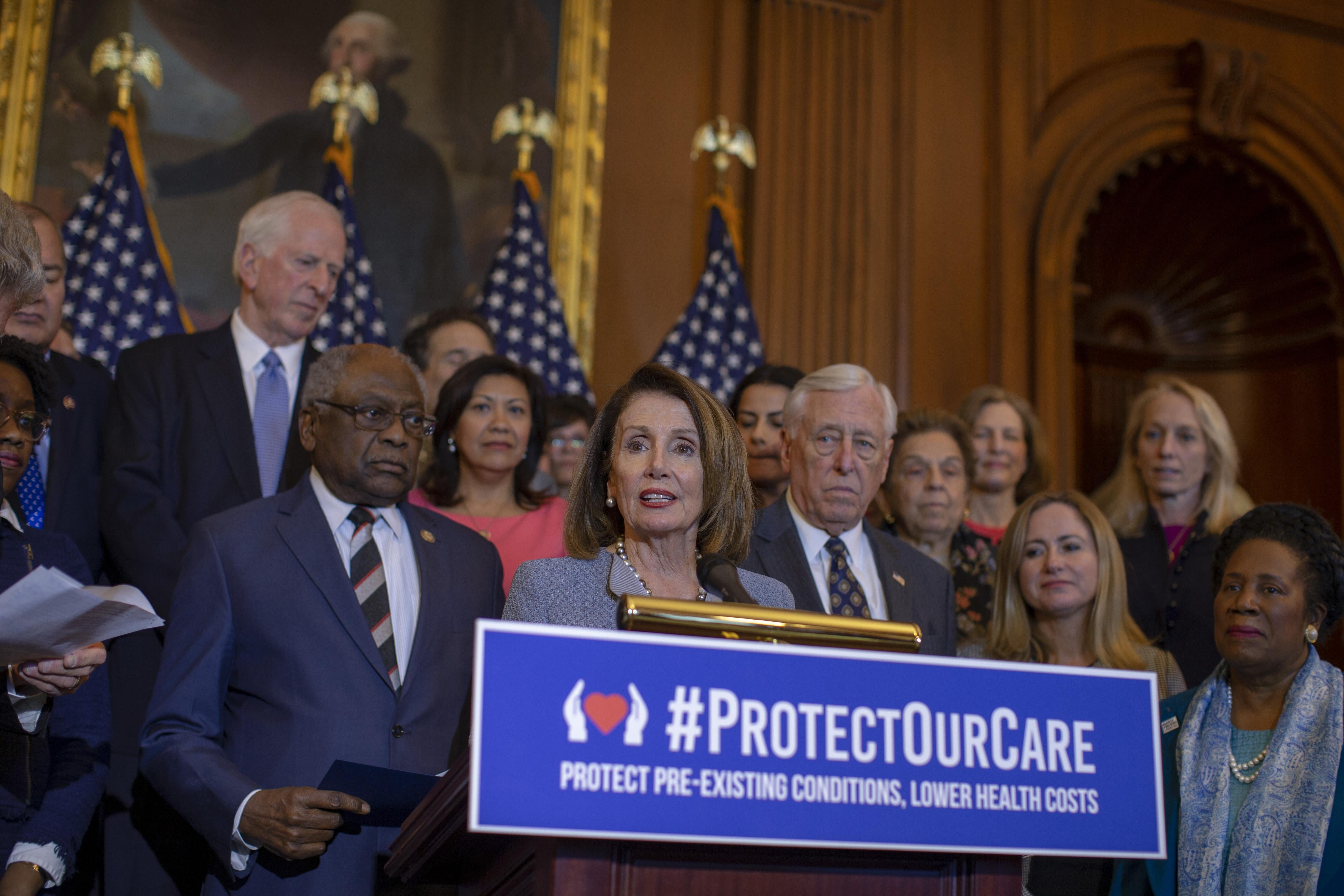 Pelosi, flanked by Steny Hoyer and other Democrats, speaks at a podium that has a "Protect Our Care" sign on it.
