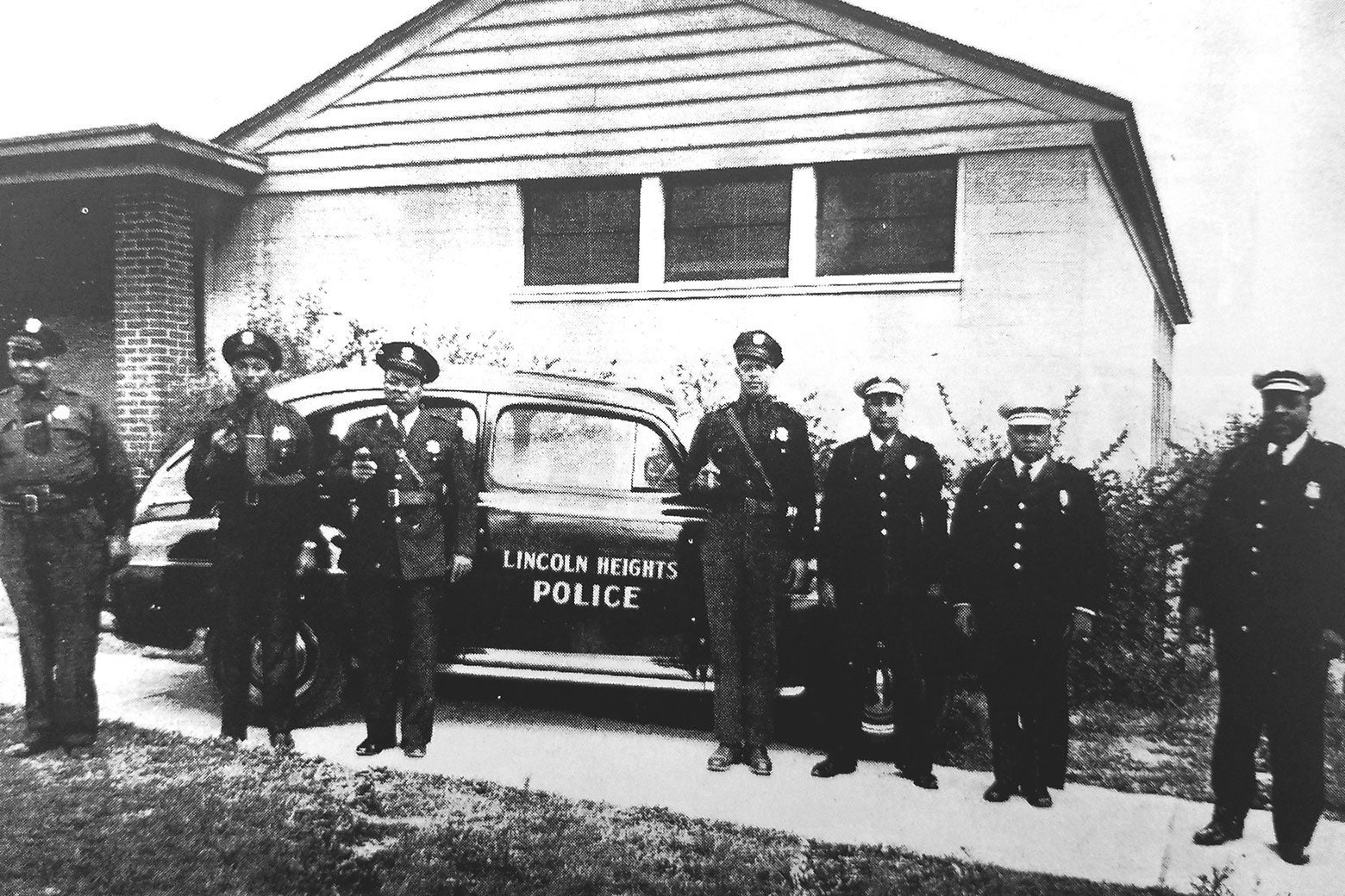 Seven men in police uniforms stand in front of a low building and a car with "Lincoln Heights Police" written on its door.