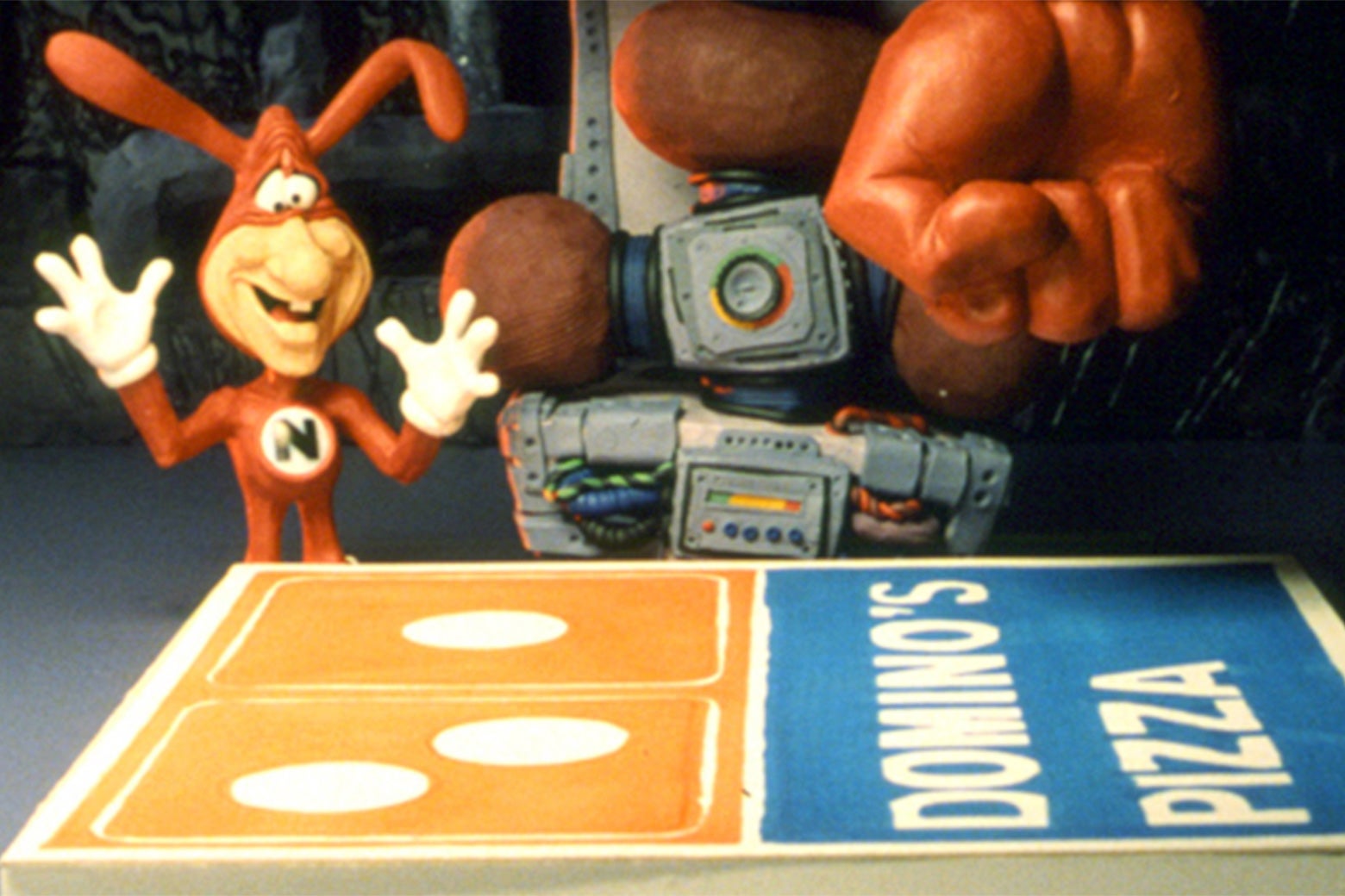The Domino's noid in a 1980s ad. The noid is a tiny humanoid creature dressed in a red Spandex suit. It has a mask and red bunny ears that are also part of the suit. It has a human face with a big nose and wears white gloves. It stands over a Dominos Pizza box. A gloved human hand hovers off-screen at the top.
