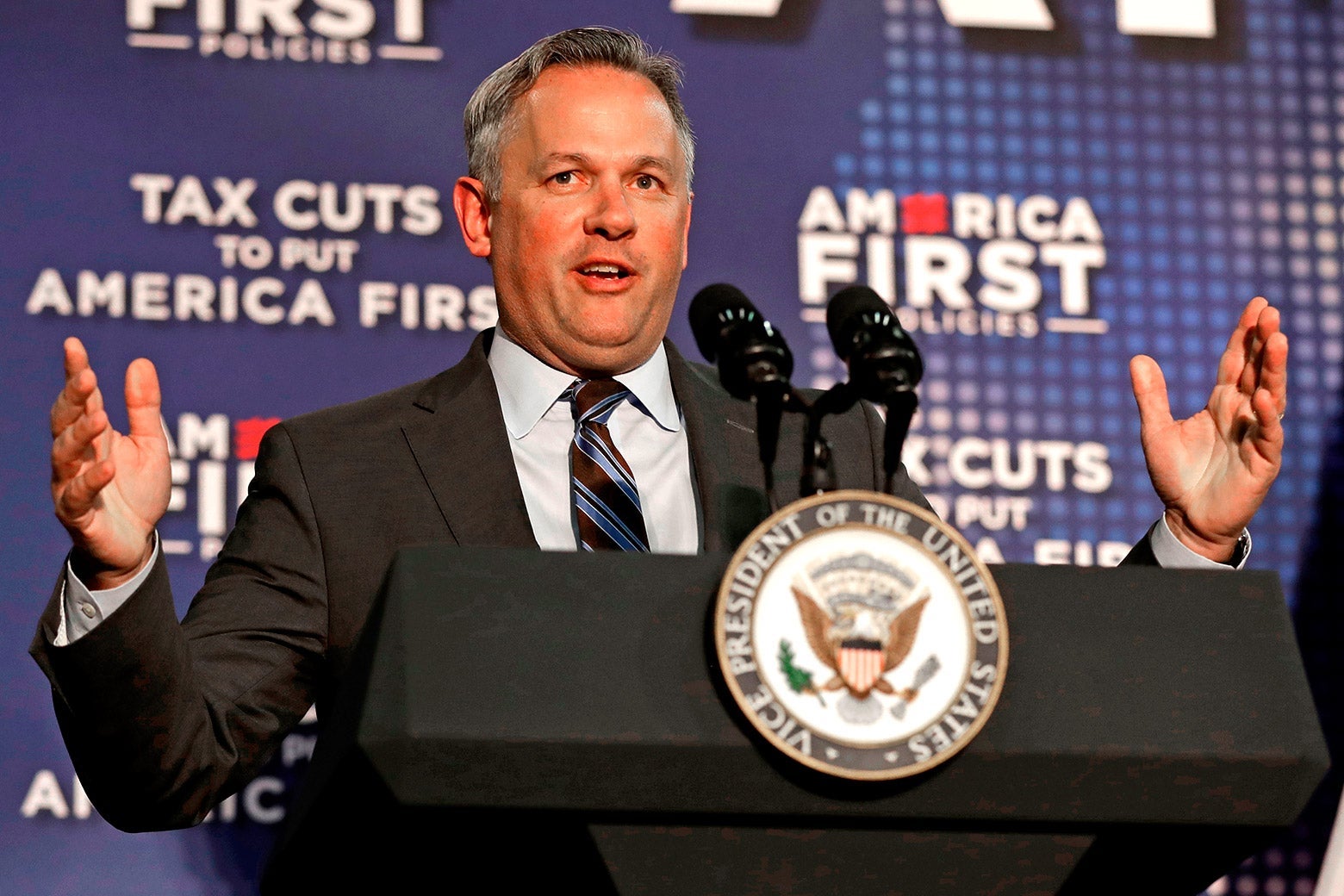 Forest raises his arms while speaking at a podium, with America First signage behind him.