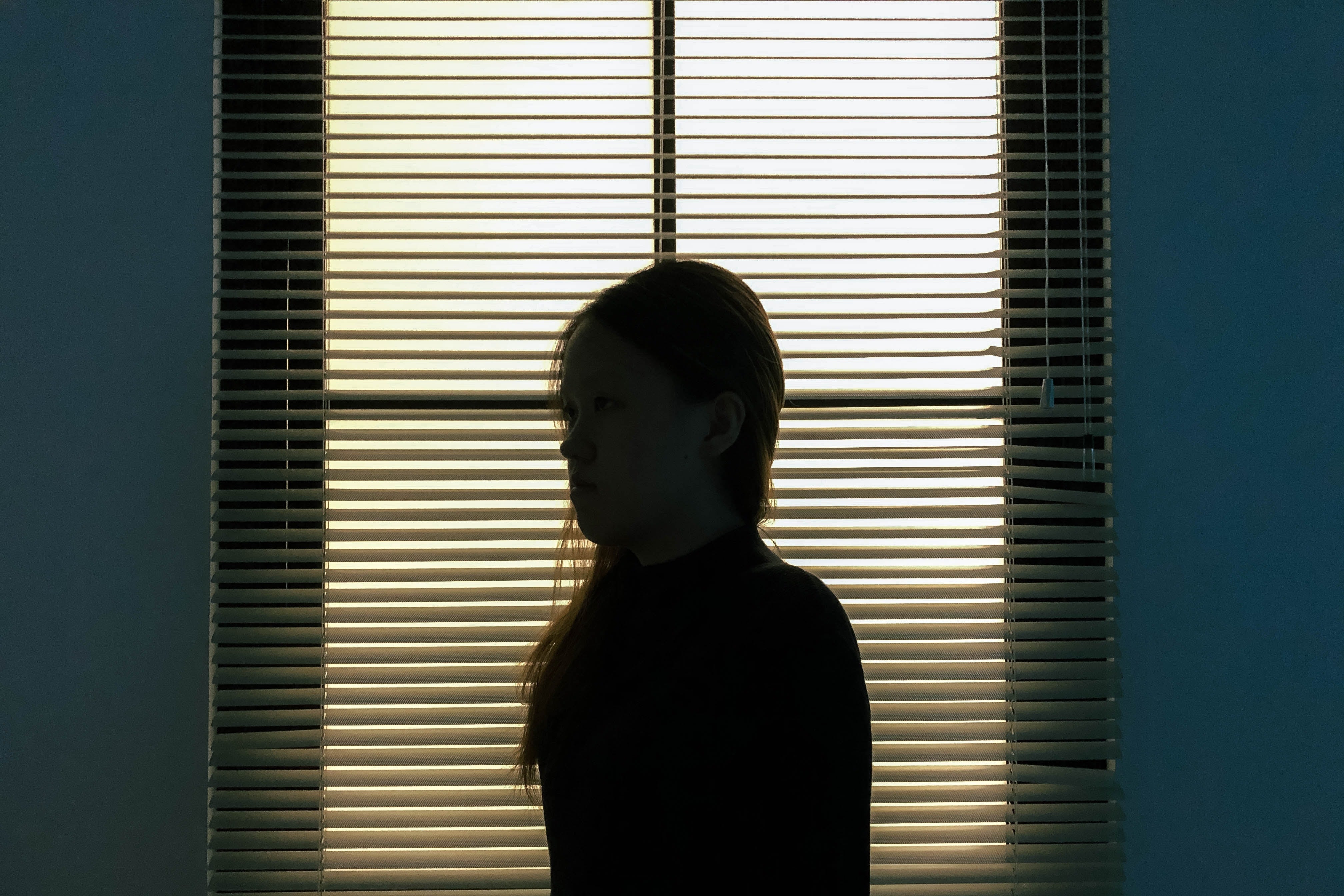 The silhouette of a woman standing in front of a window with the blinds closed.