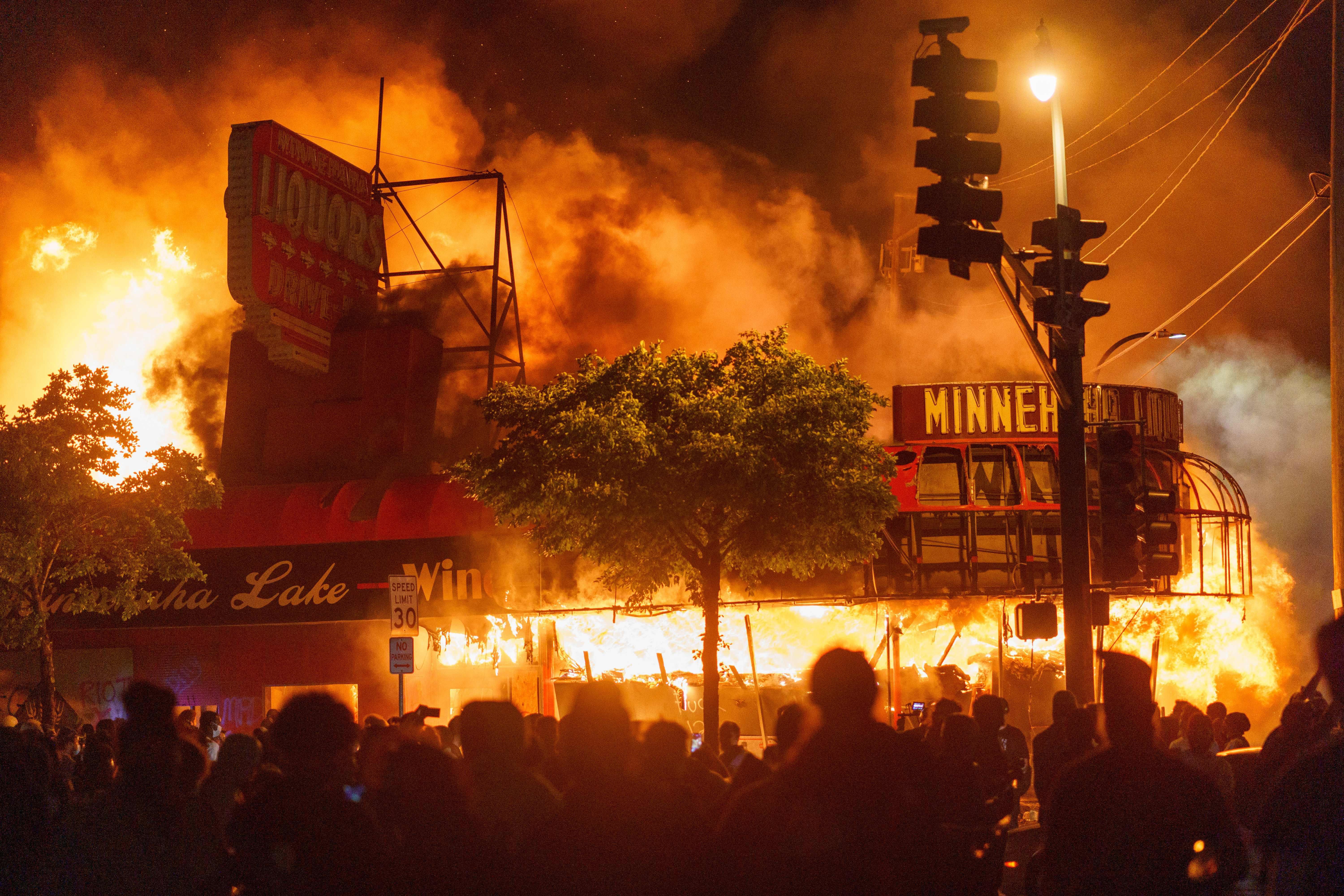 Protesters gather in the street in front of a burning liquor store. Smoke fills the night sky.