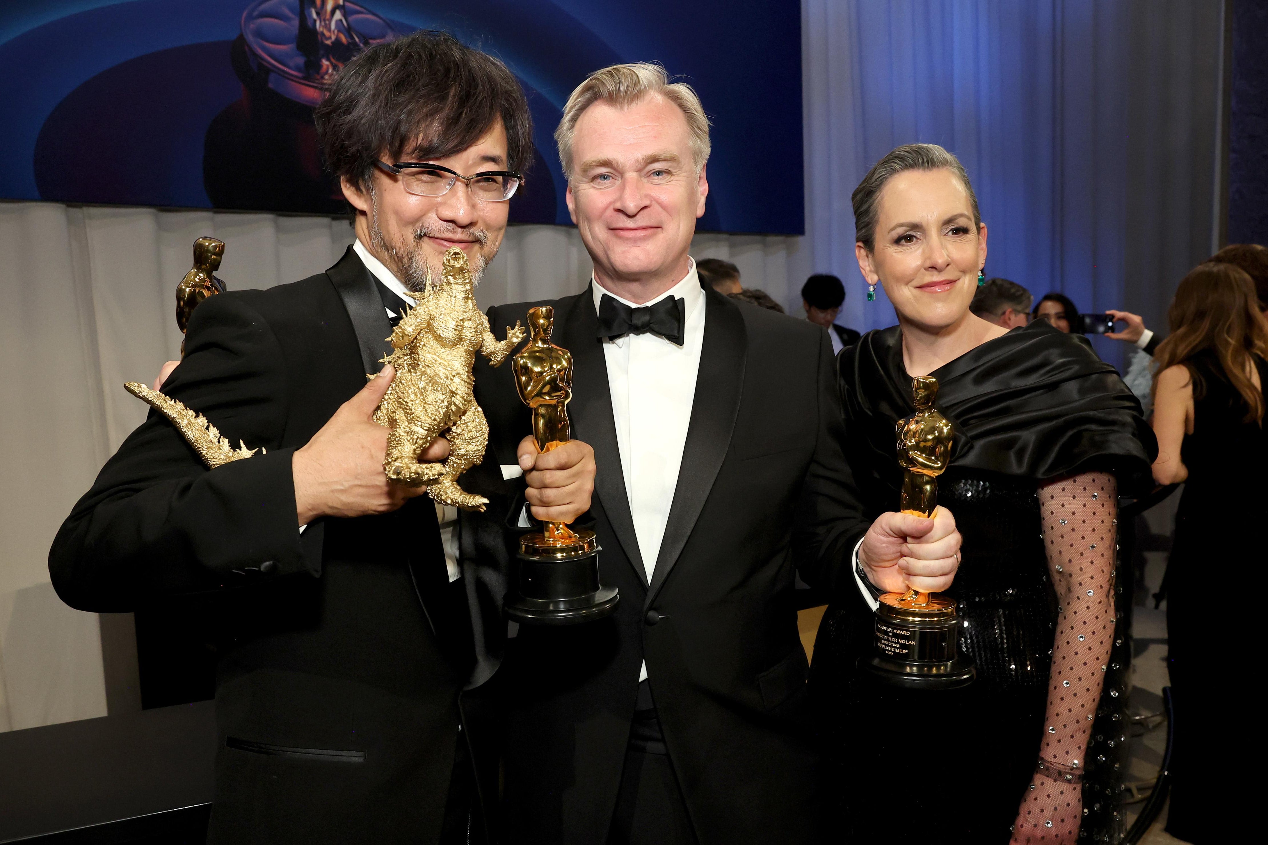 Takashi Yamazaki, winner of the Best Visual Effects award for “Godzilla Minus One,” and Christopher Nolan and Emma Thomas, winners of the Best Picture award for 'Oppenheimer,' huddle together while smiling wide and holding up their trophies. Yamazaki also holds up a little golden Godzilla.