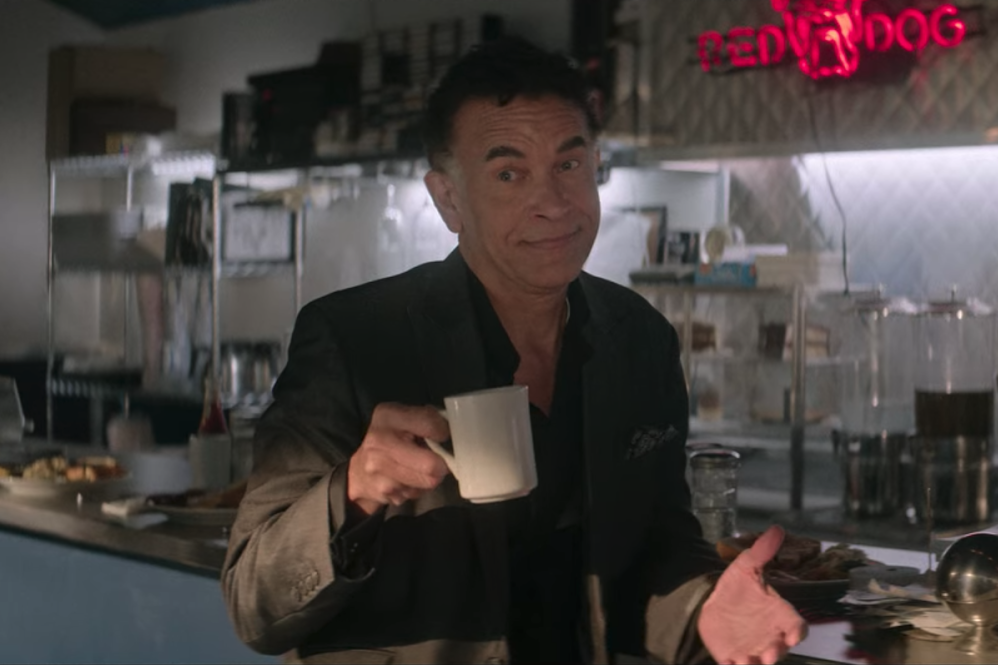 Brian Stokes Mitchell sits at a diner counter with a coffee mug in hand, his hands out in a shoulder shrugging motion.