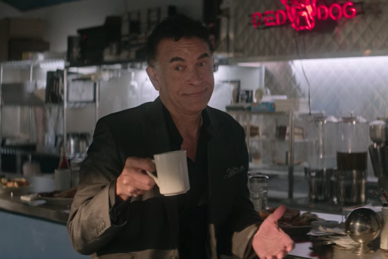 Brian Stokes Mitchell sits at a diner counter with a coffee mug in hand, his hands out in a shoulder shrugging motion.