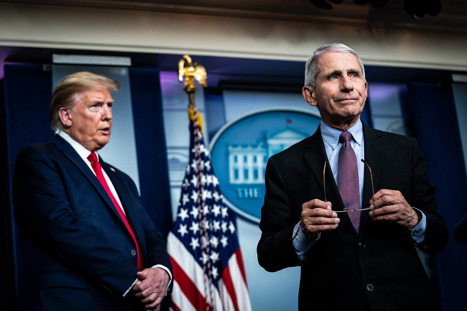 Trump stands back and to the side as Fauci stands forward holding his glasses.