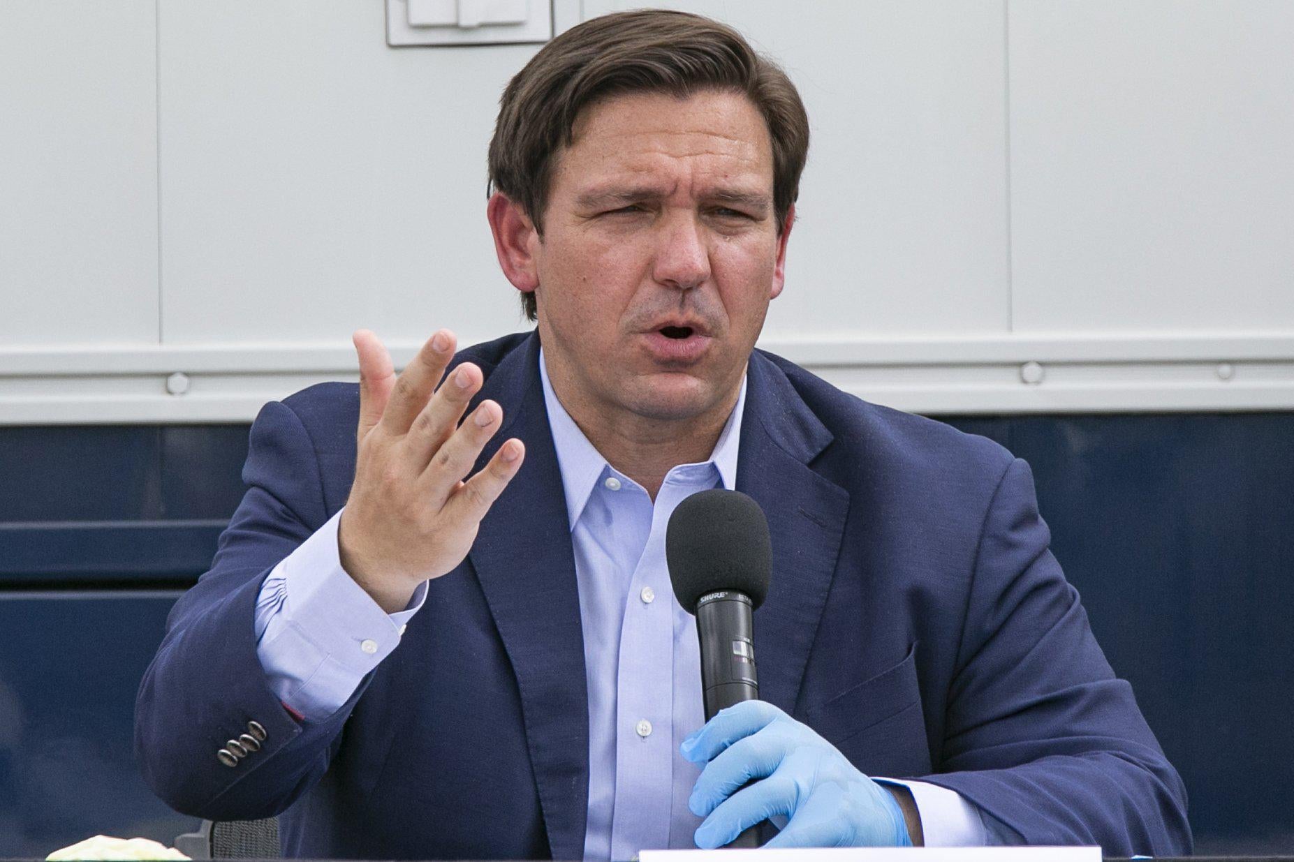 Ron DeSantis speaks and holds a mic with a gloved hand.