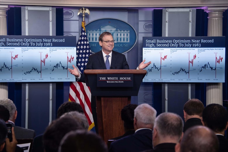 Kevin Hassett stands at briefing room podium flanked by large posters of economic graphs.