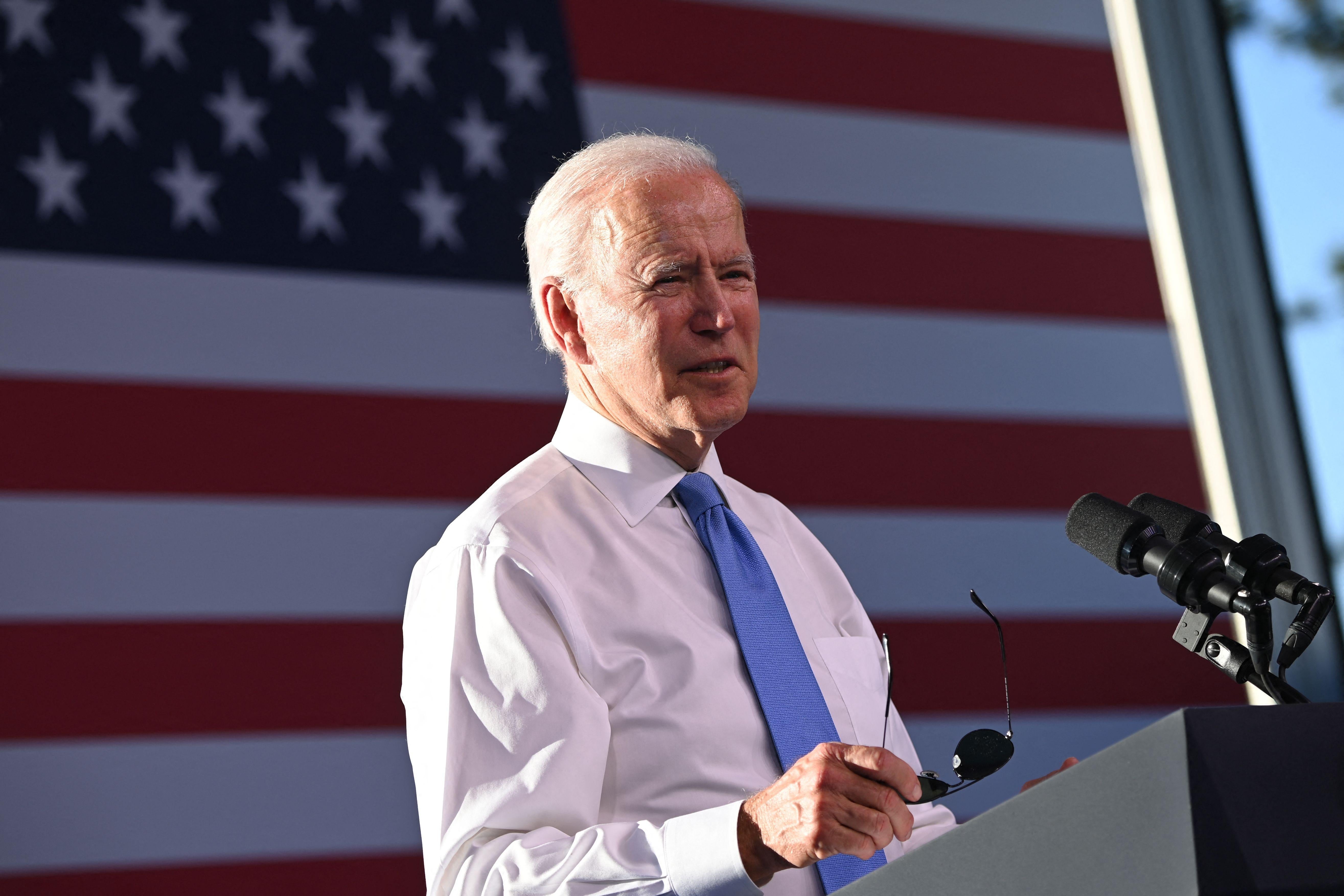 Joe Biden standing at a lectern in front of an American flag, holding his aviator sunglasses.