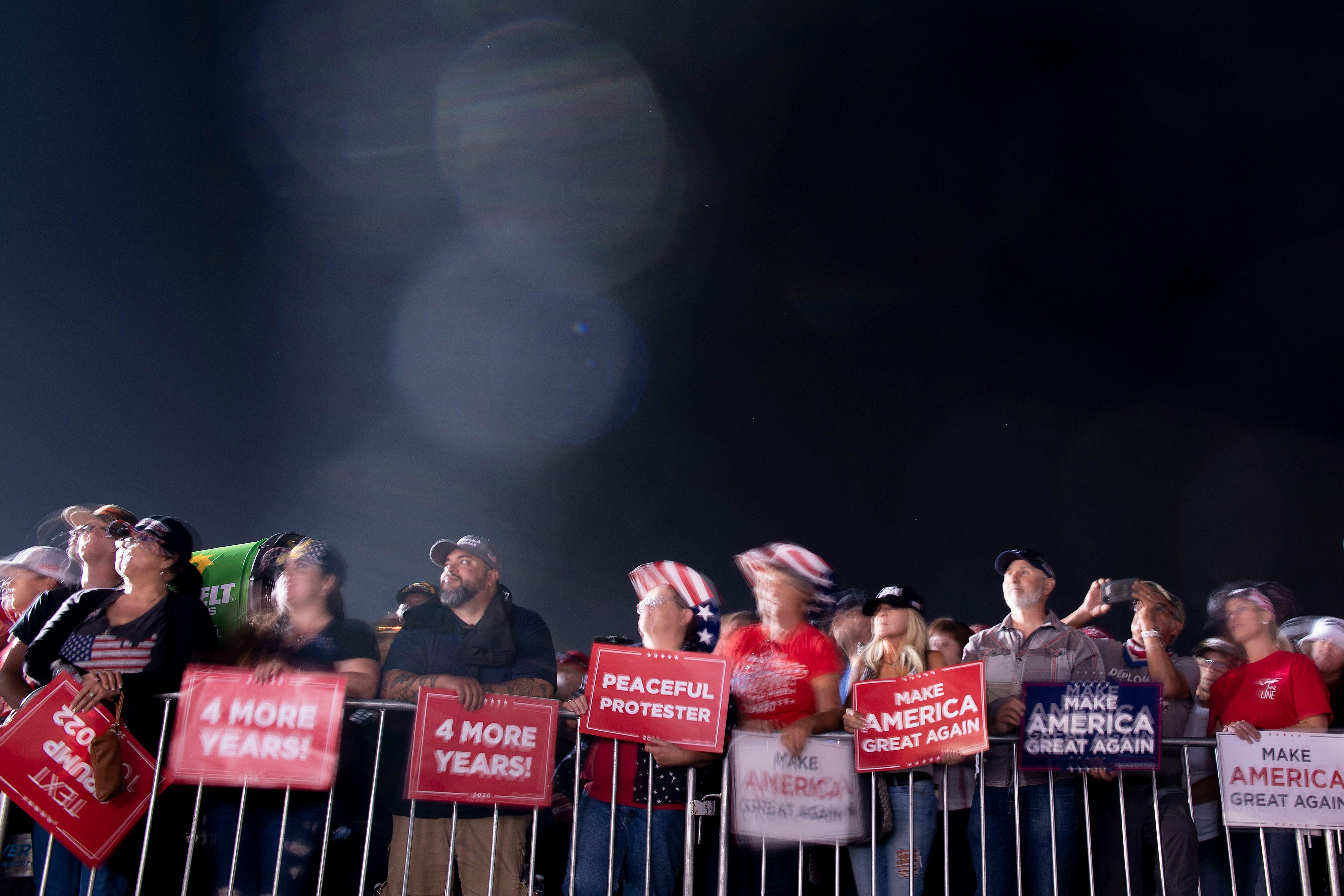 A crowd of Trump supporters behind a barrier at night. They are holding signs that say "Four More Years" and "Make America Great Again."