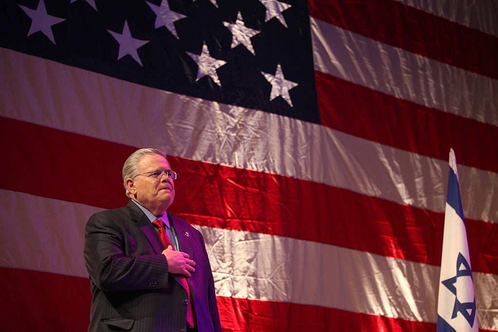 John Hagee standing with his hand over his heart in front of an American-flag backdrop with an Israeli flag to his left.