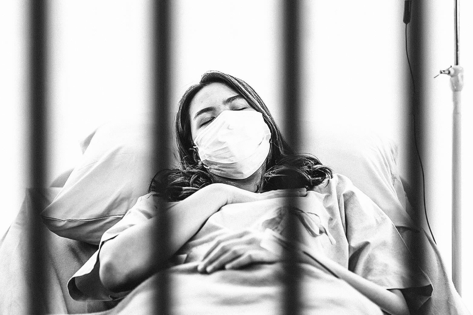A woman lying on a hospital bed behind bars and wearing a face mask