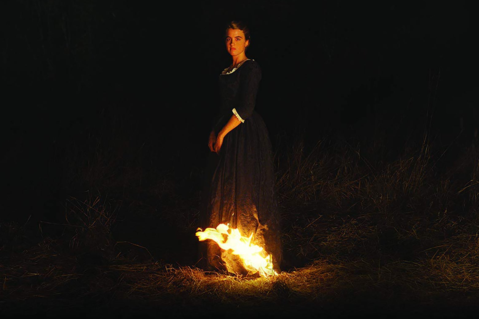 A still from Portrait of a Lady on Fire. It's a night shot, with intense chiaroscuro; Adèle Haenel stands in the center, looking at the camera with a neutral impression. Her long dress is on fire at the hem, and her face is illuminated by the fire. 