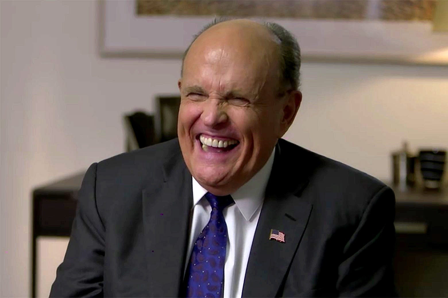 Rudy Giuliani in a medium shot, sitting in a hotel room laughing.