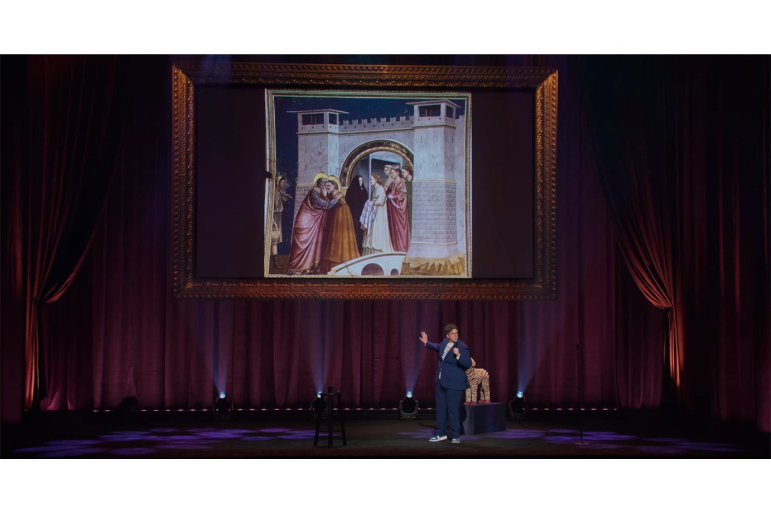 Hannah Gadsby onstage in front of a projection of Giotto's Meeting at the Golden Gate, which shows two people in brightly colored clothes embracing; a woman dressed in black is walking away from them.