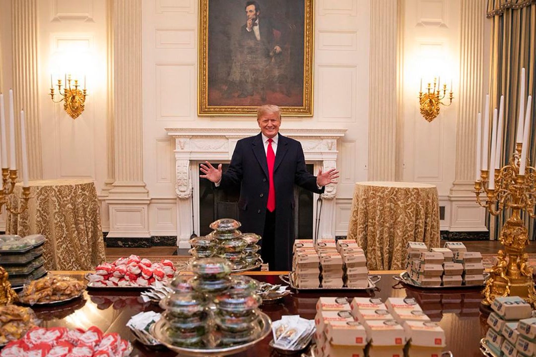 Donald Trump stands leeringly before a banquet table stacked with McDonald's burgers, Chick-Fil-A sandwiches, and other fast food items in a White House ballroom.