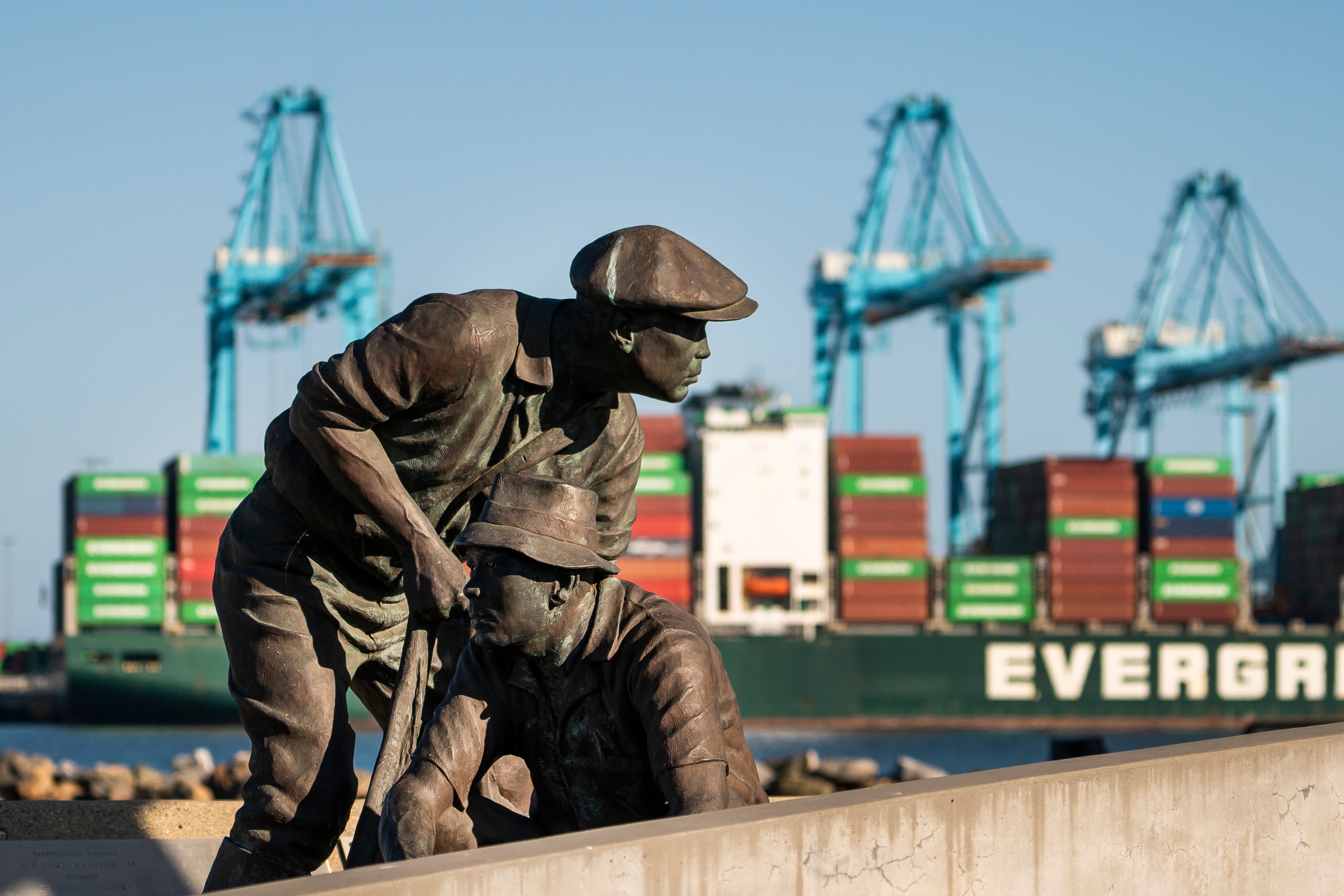 A statue of two fishermen is in front of distant shipping containers and cranes.