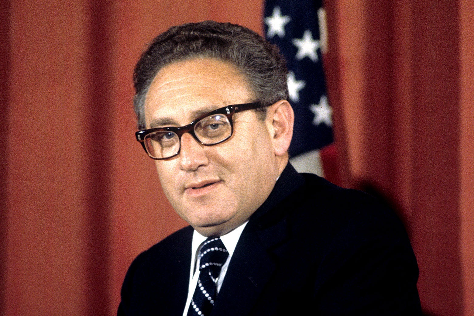 Kissinger in a black suit and tie smirking.