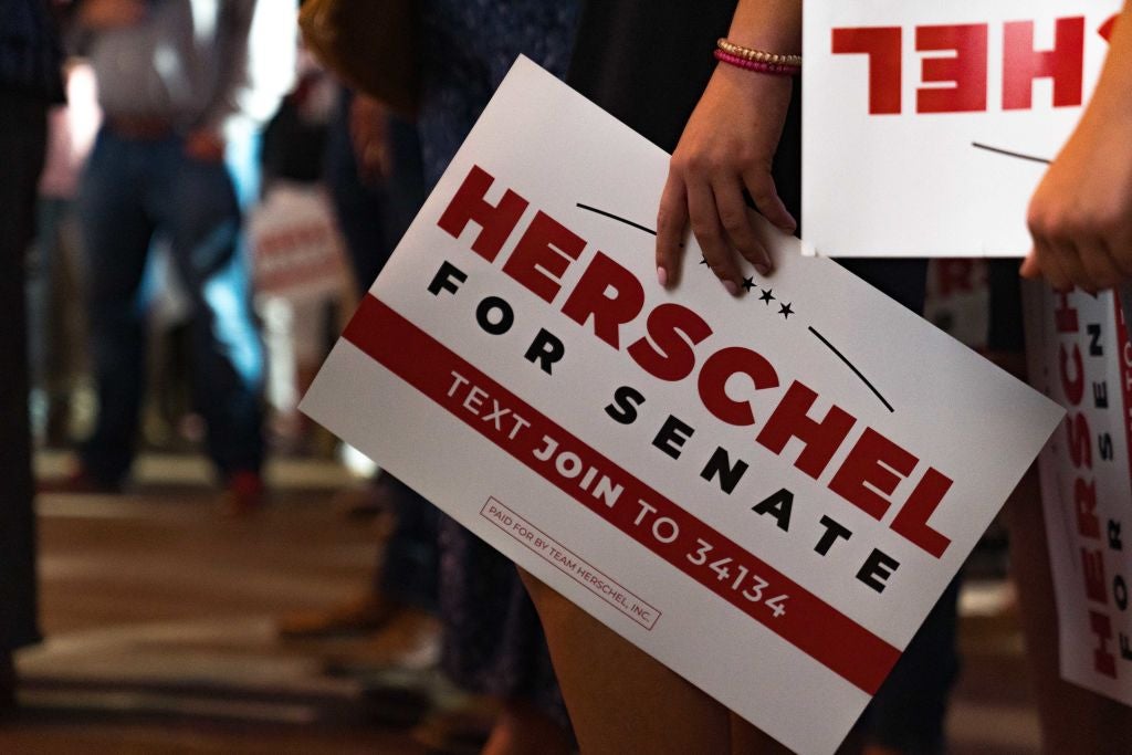 A rallygoer holds a "Herschel for Senate" sign in a shot taken at waist level.