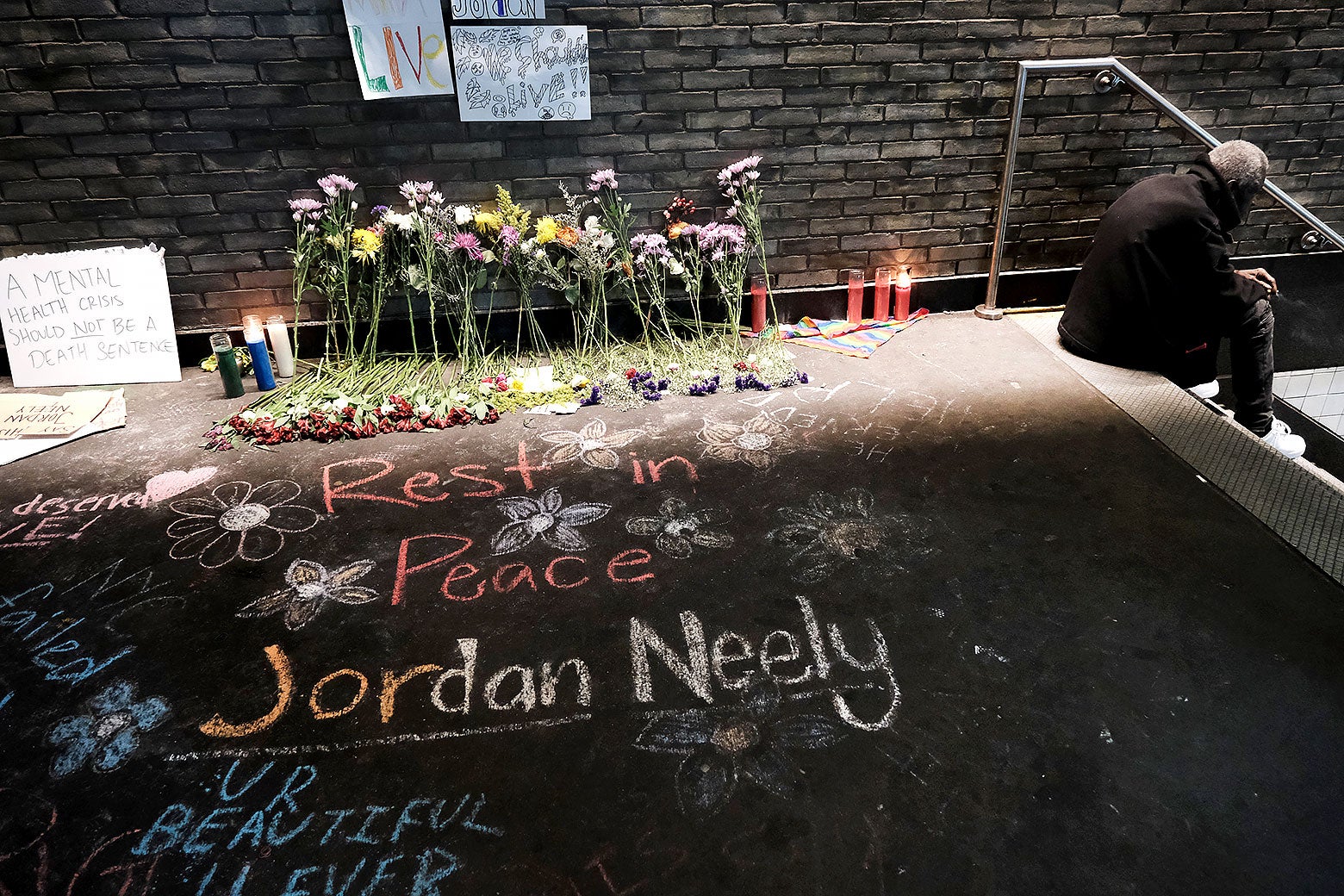 A brick exterior with flowers and prayer candles, with "Rest in Peace Jordan Neely" written in chalk on the concrete.
