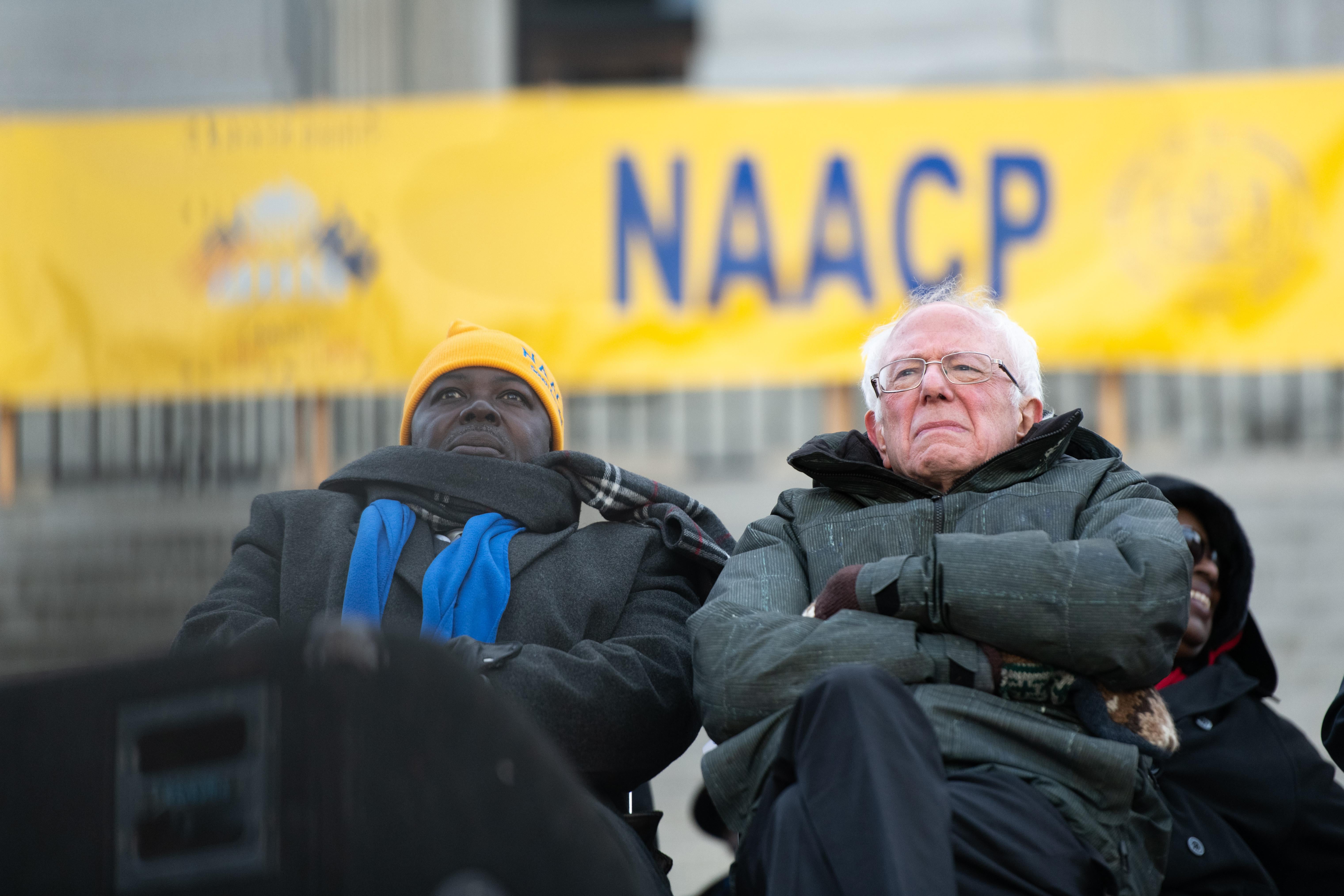 Bernie Sanders stands in front of an NAACP banner, arms crossed.