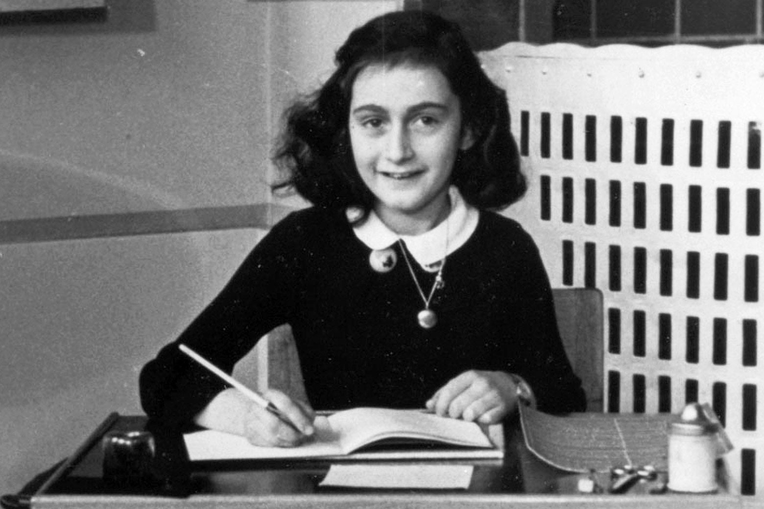 Photo of Anne Frank sitting at a school desk and holding a pencil to an open notebook page.