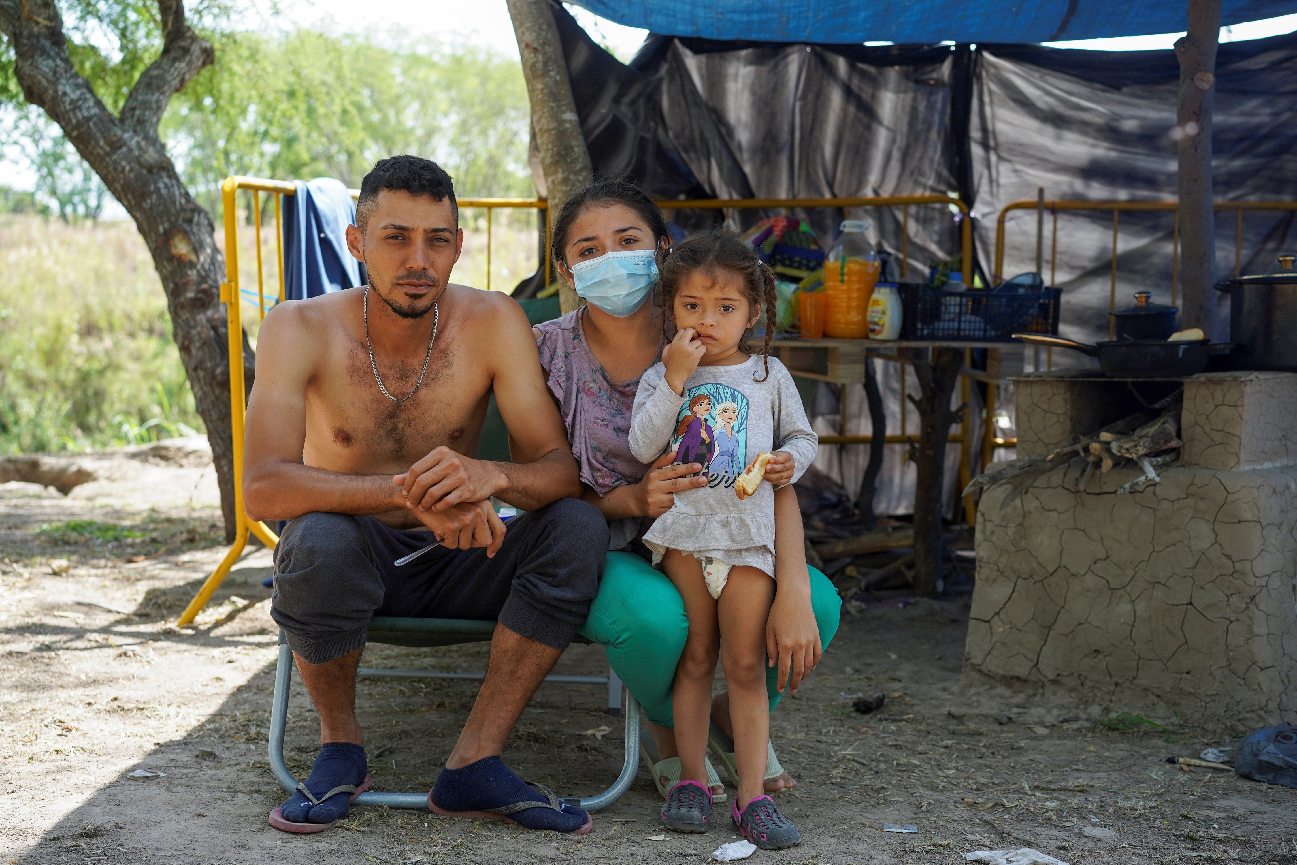 A shirtless man, a woman in a mask, and a small girl in a diaper pose for a photo in front of a makeshift tent.
