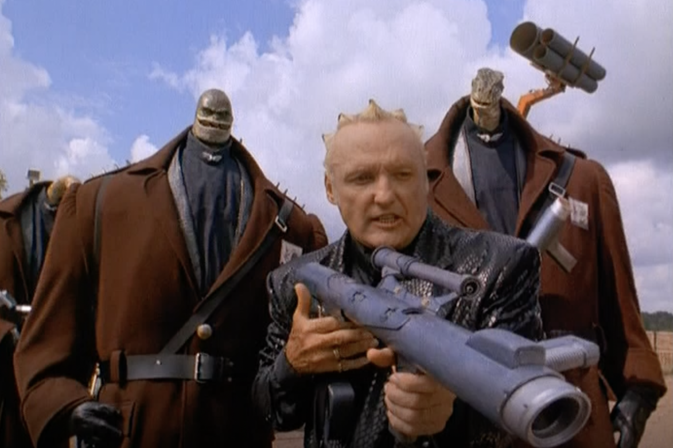 two very large men with tiny lizard heads on either side of dennis hopper, who is holding a bazooka