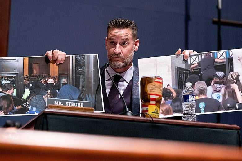 Steube stands at a podium holding up two photos. He raises both eyebrows in a questioning look.