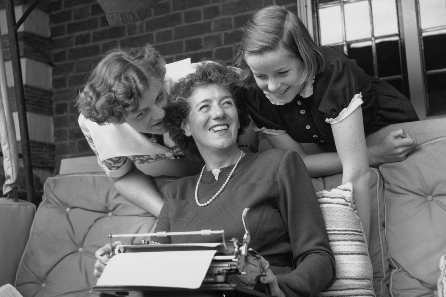 Two young teenage girls lean affectionately over a woman seated with a typewriter in her lap. 