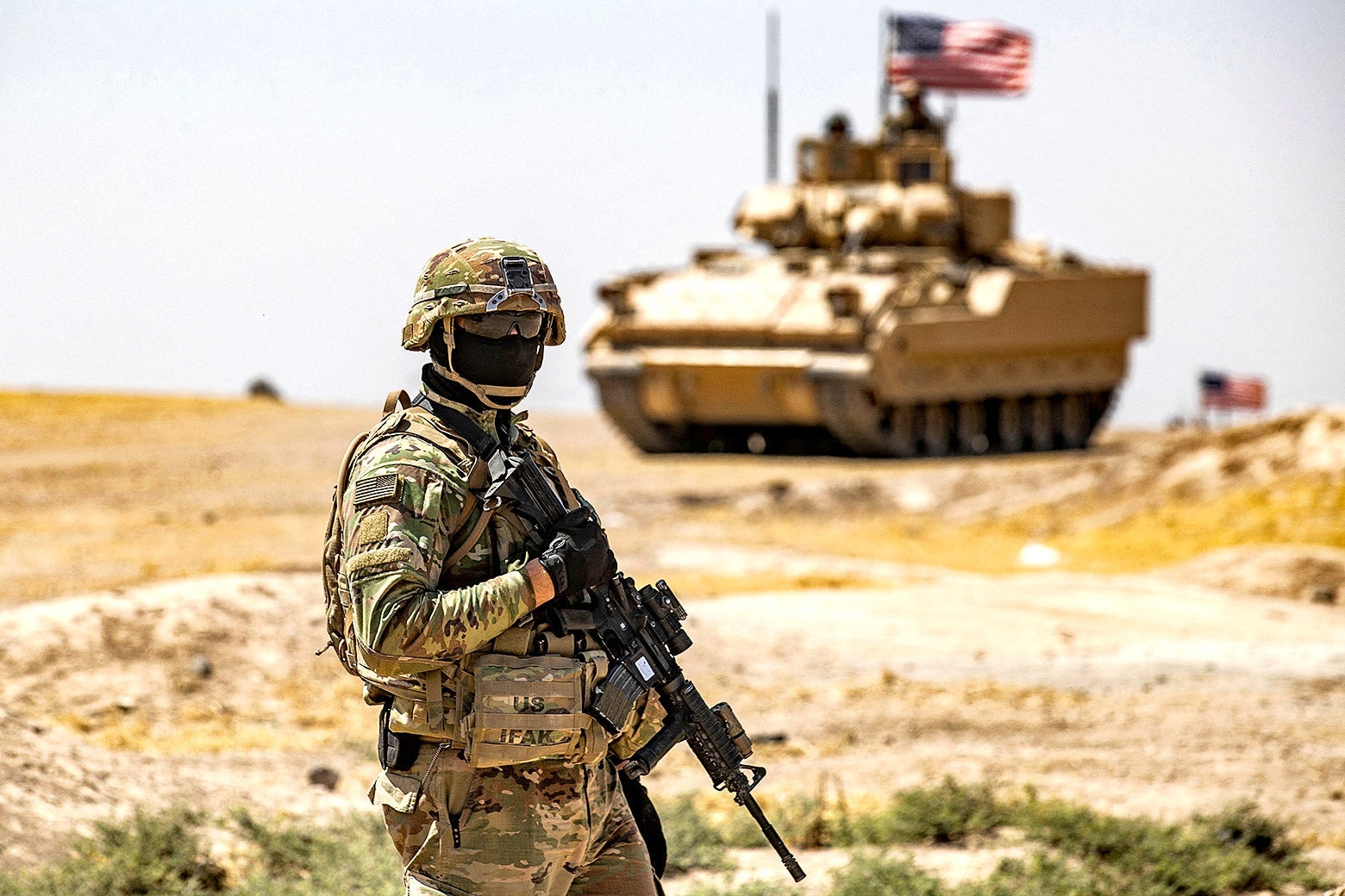 An American soldier wearing a black mask and holding a gun stands in a desert landscape with an American tank in the distance
