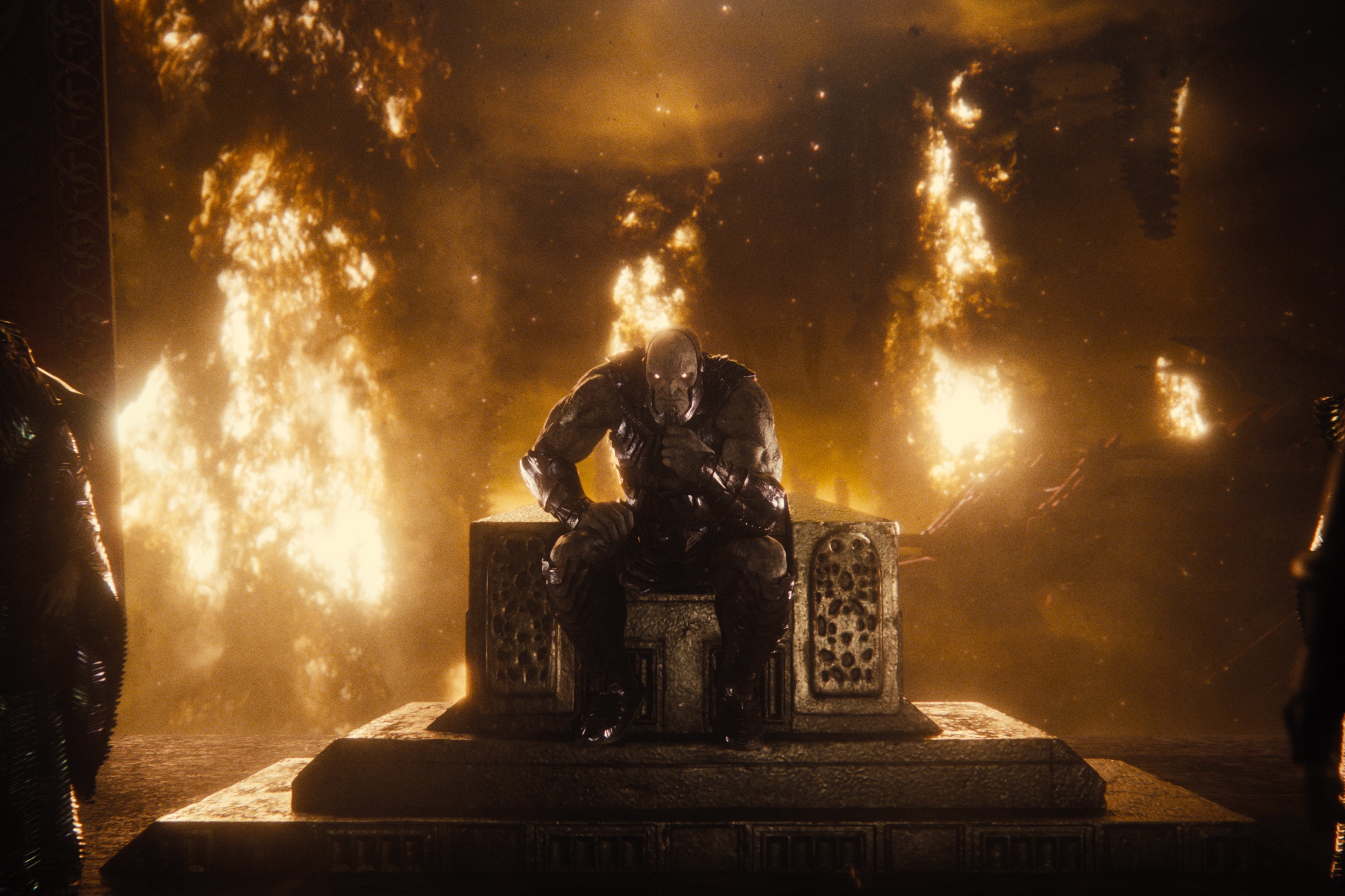 A masked figure sits on a throne in front of a flaming backdrop.