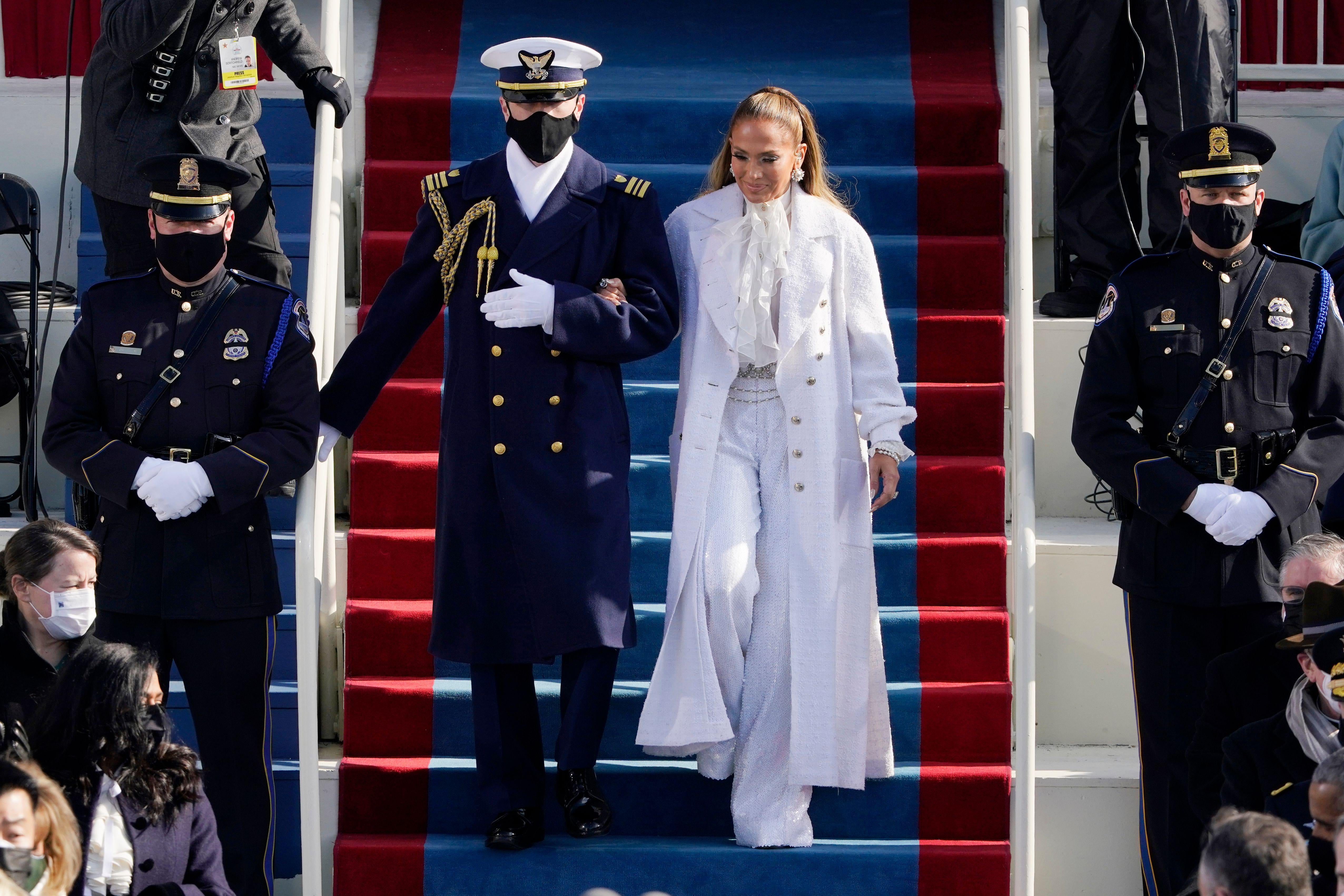 Jennifer Lopez arrives escorted by a member of the military. She wears an all-white outfit with wide-legged pants and a flowy blouse under a long white jacket.