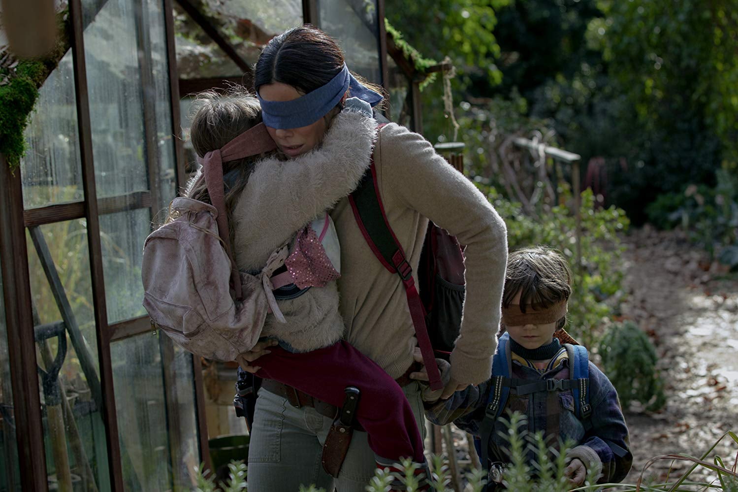 A woman carries a small girl through the forest as a young boy walks alongside them. They are all blindfolded. 