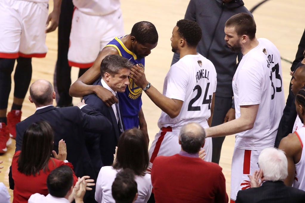 Kevin Durant walks past Toronto players while supported by a trainer.