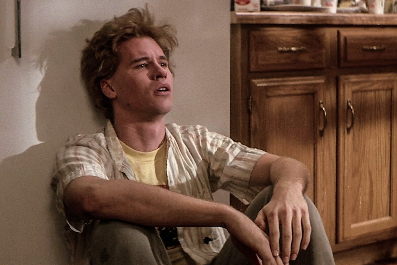Chris Knight, the character played by Val Kilmer in Real Genius, sits leaning against a wall, with an exasperated expression on his face. Behind him to the right are wood cabinets and a countertop. 