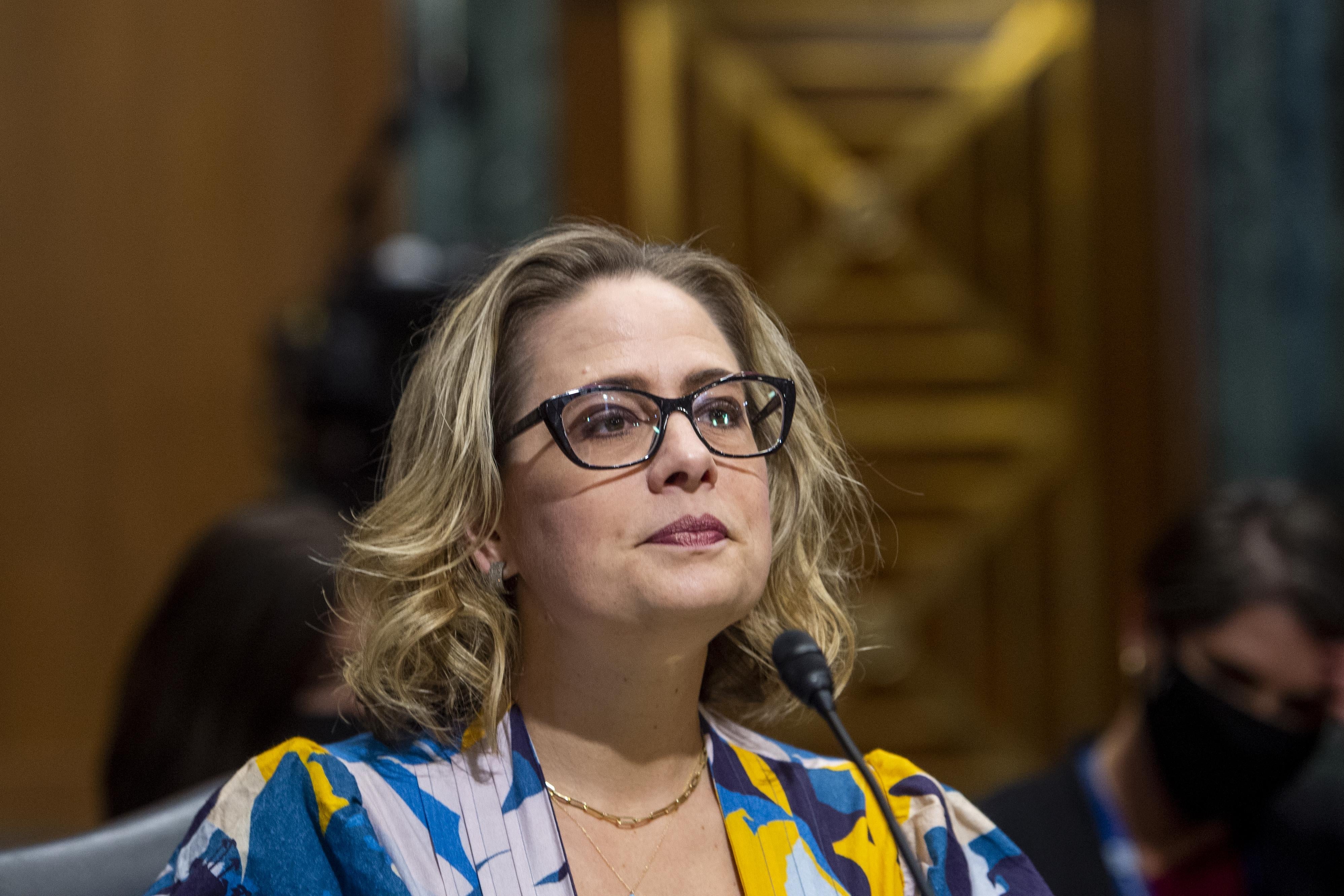 Kyrsten Sinema purses her lips while sitting in front of a mic in a hearing room