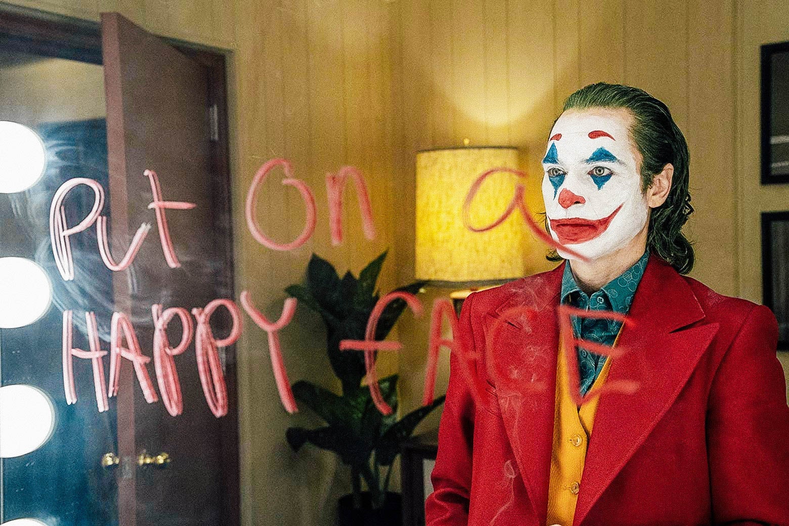 In this still of Joaquin Phoenix in Joker, Phoenix is clad in clown makeup and looking at a green room mirror with the words "Put on a happy face" written on it in marker or makeup.