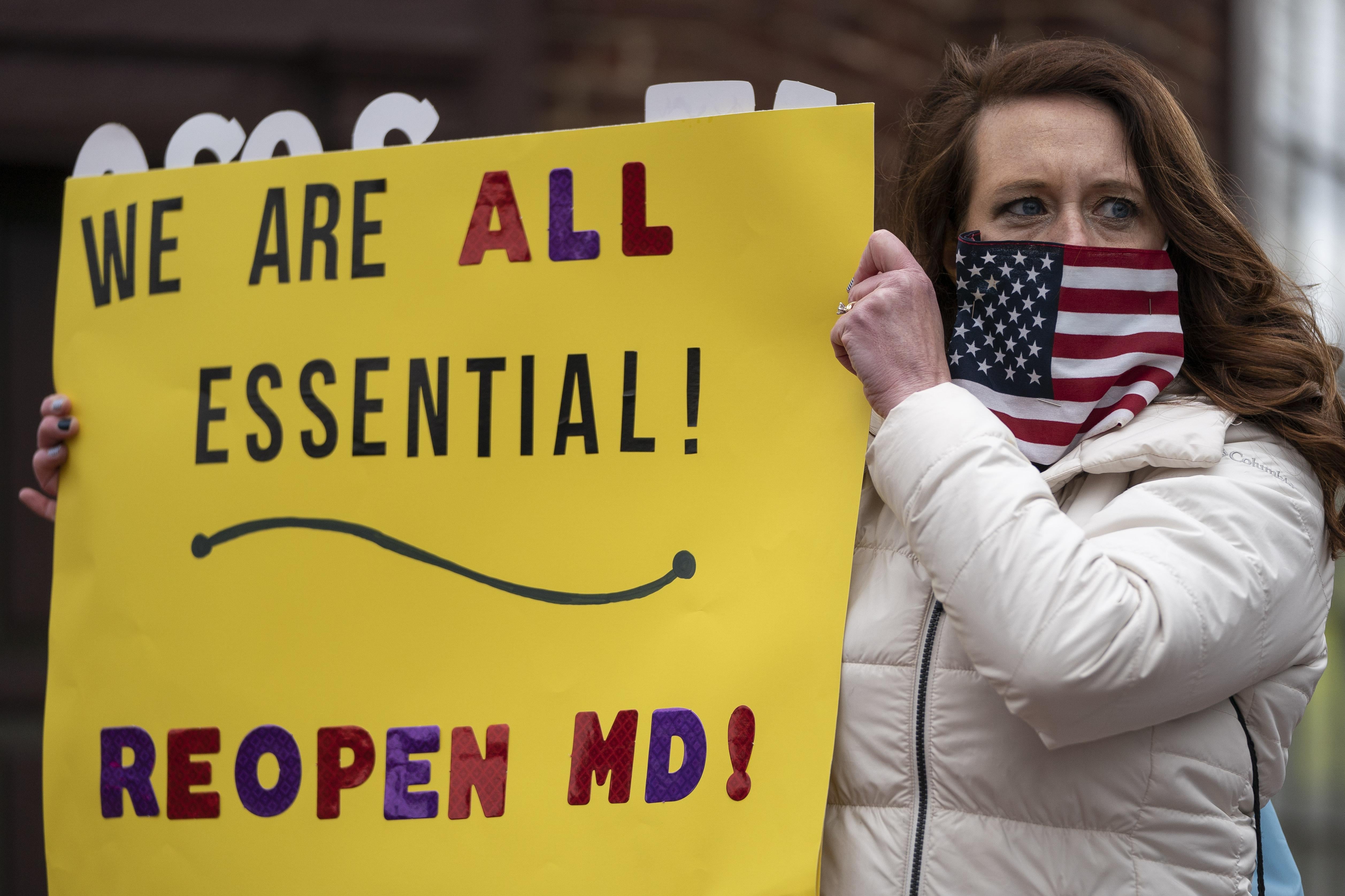 A protester wearing an American flag face covering holds up a sign that says "We are all essential! Reopen MD!"