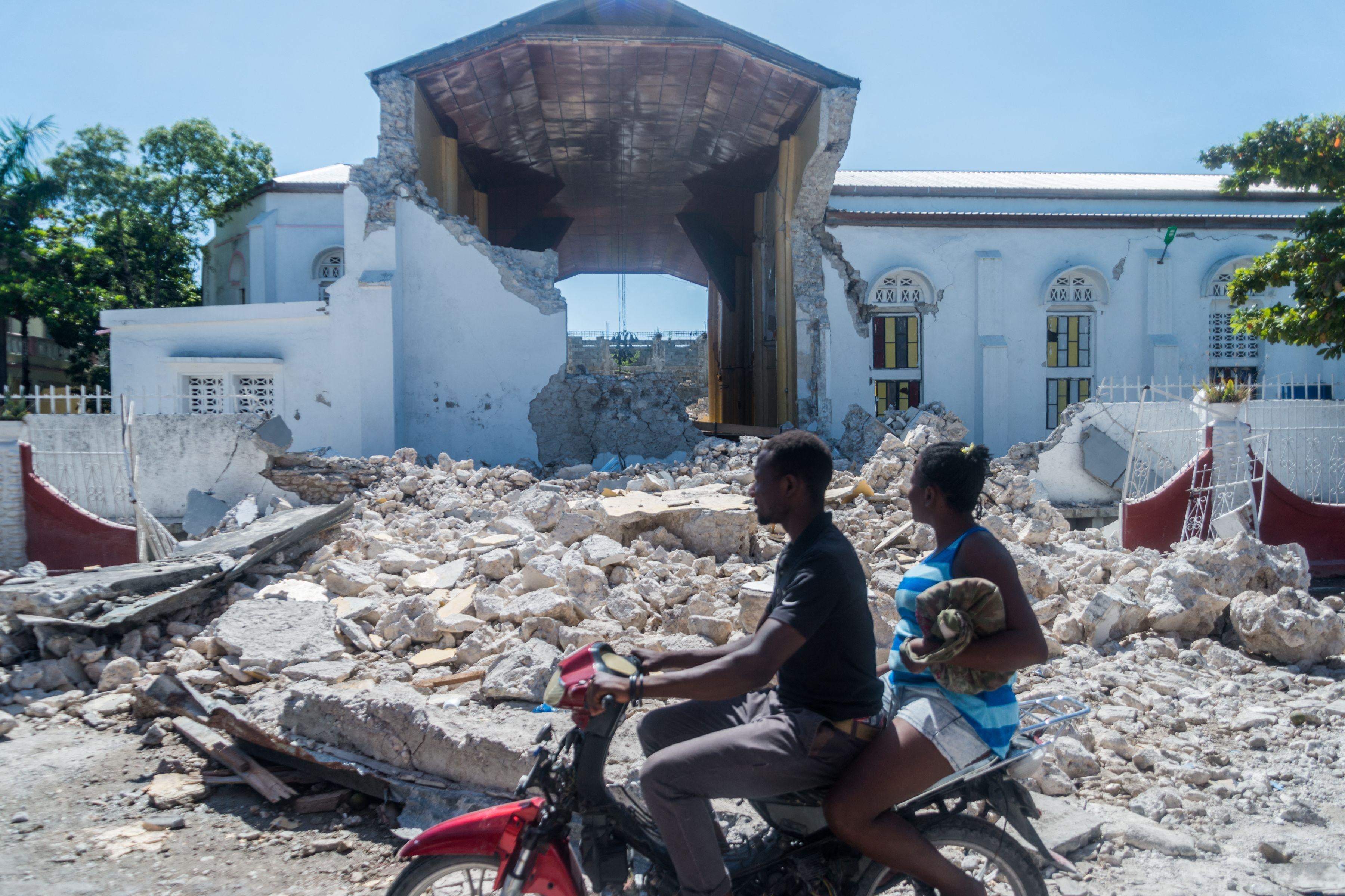 Two people on a motorbike ride past a pile of rubble in front of the husk of the church building