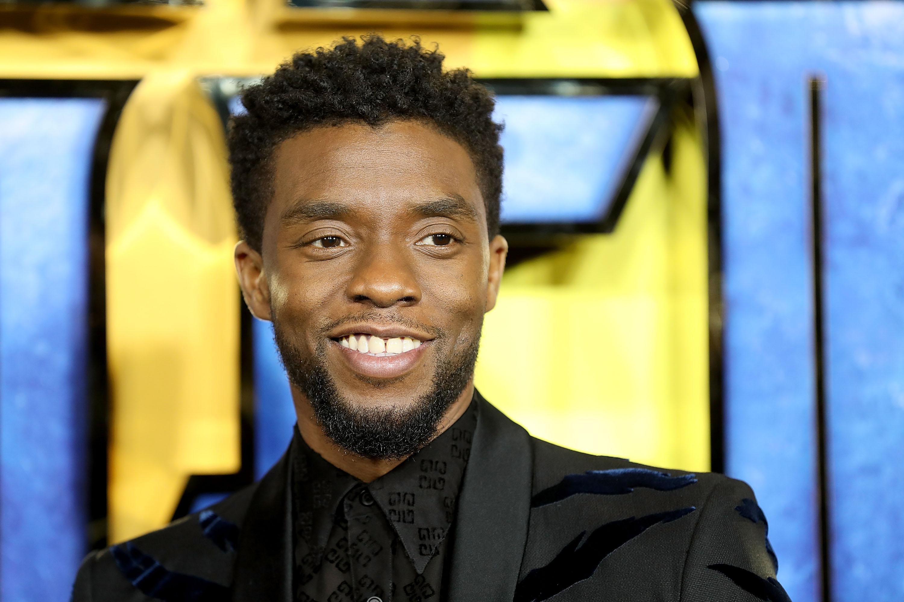 Actor Chadwick Boseman wearing a black suit in front of a blue curtain, smiling.