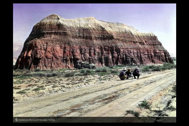 Color photo of three men crouched on a road in front of a large rock outcrop with a car parked at its base