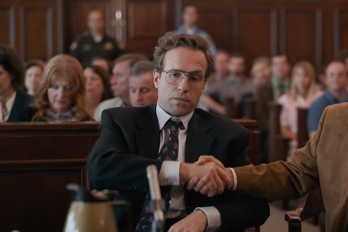 Rafe Spall sits in a courtroom, looking smug,